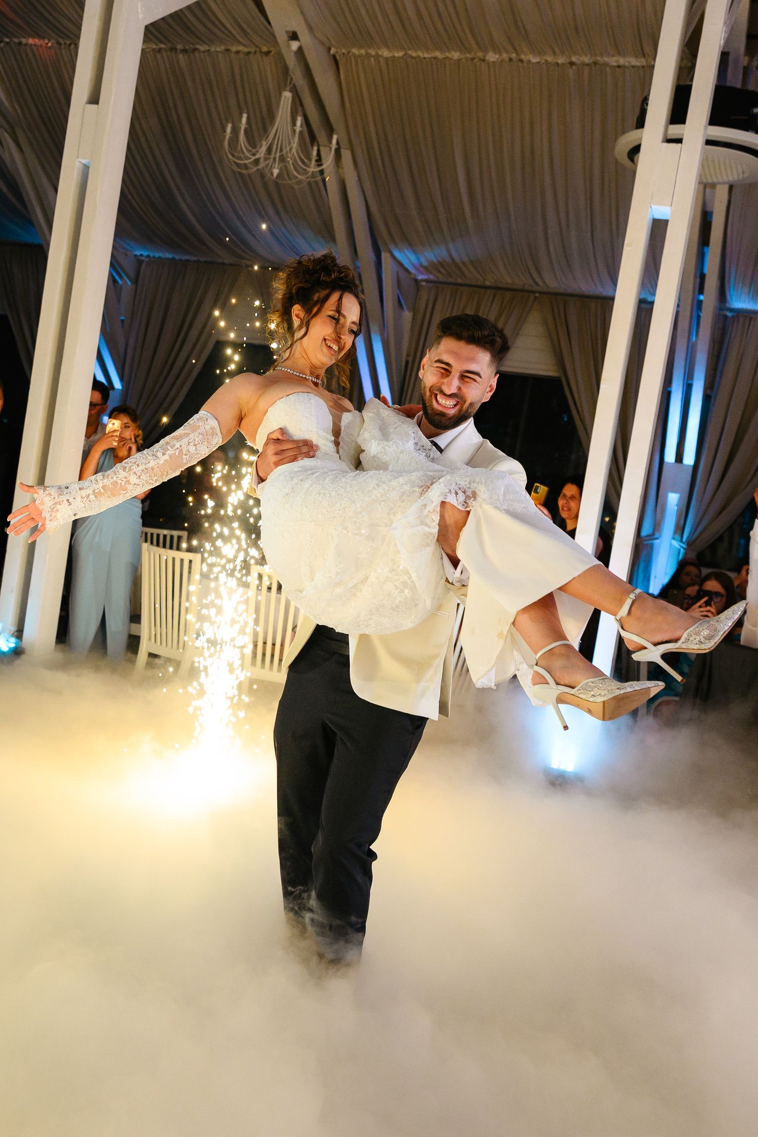 Groom carries bride on foggy dance floor with sparkling fireworks at elegant wedding reception.