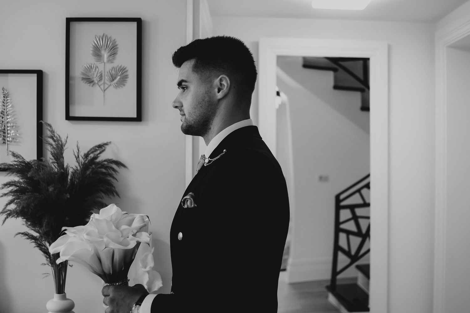 Groom in black suit holding white flowers in profile view indoors.