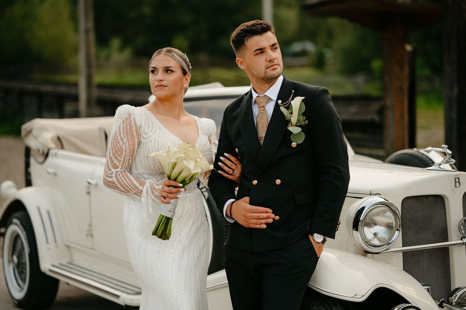 Bride in embellished gown and groom in navy suit standing before a classic white vintage car.