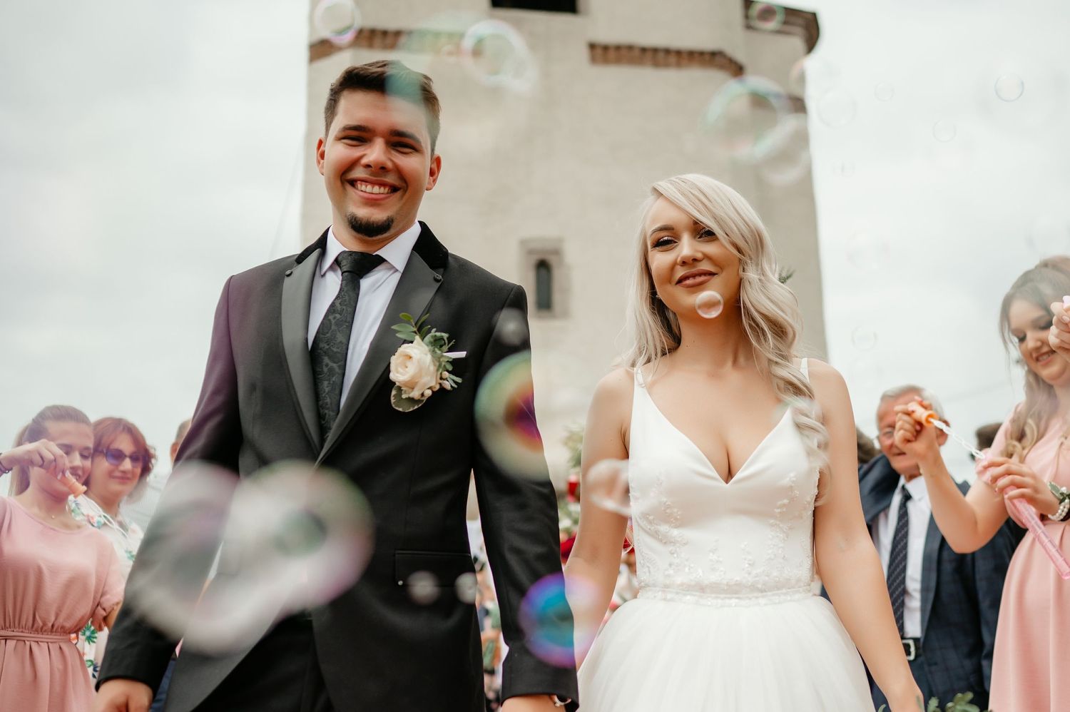 Smiling newlywed couple walks through bubbles blown by guests outside a historic stone tower.