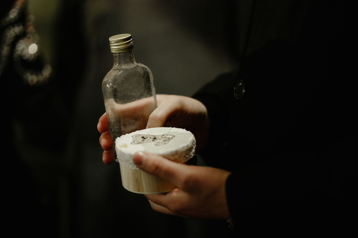 Hands holding a small glass bottle and round container at a religious baptism ceremony.