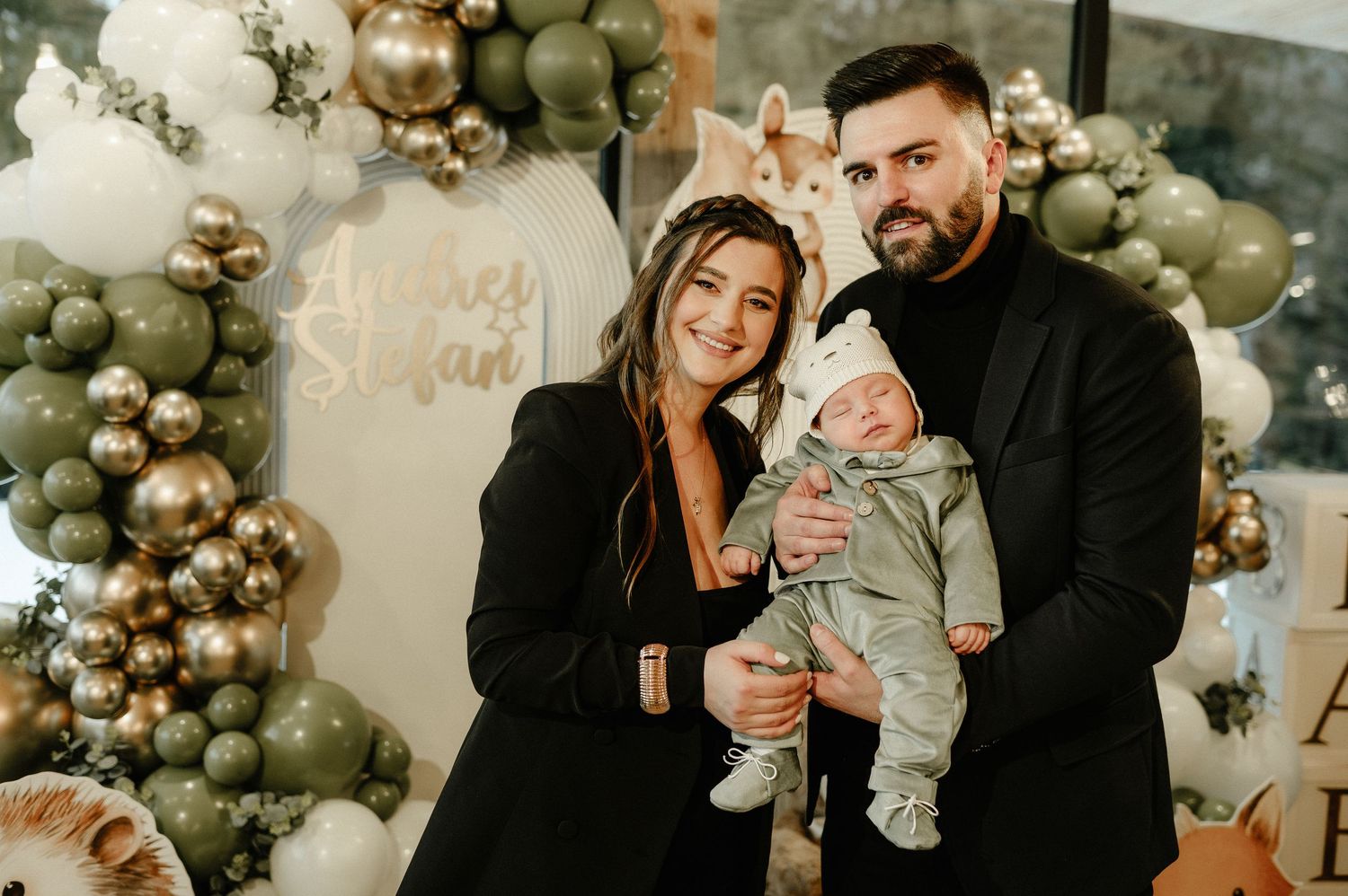 Happy couple in black outfits holding newborn baby at decorated baby shower with balloon arch.