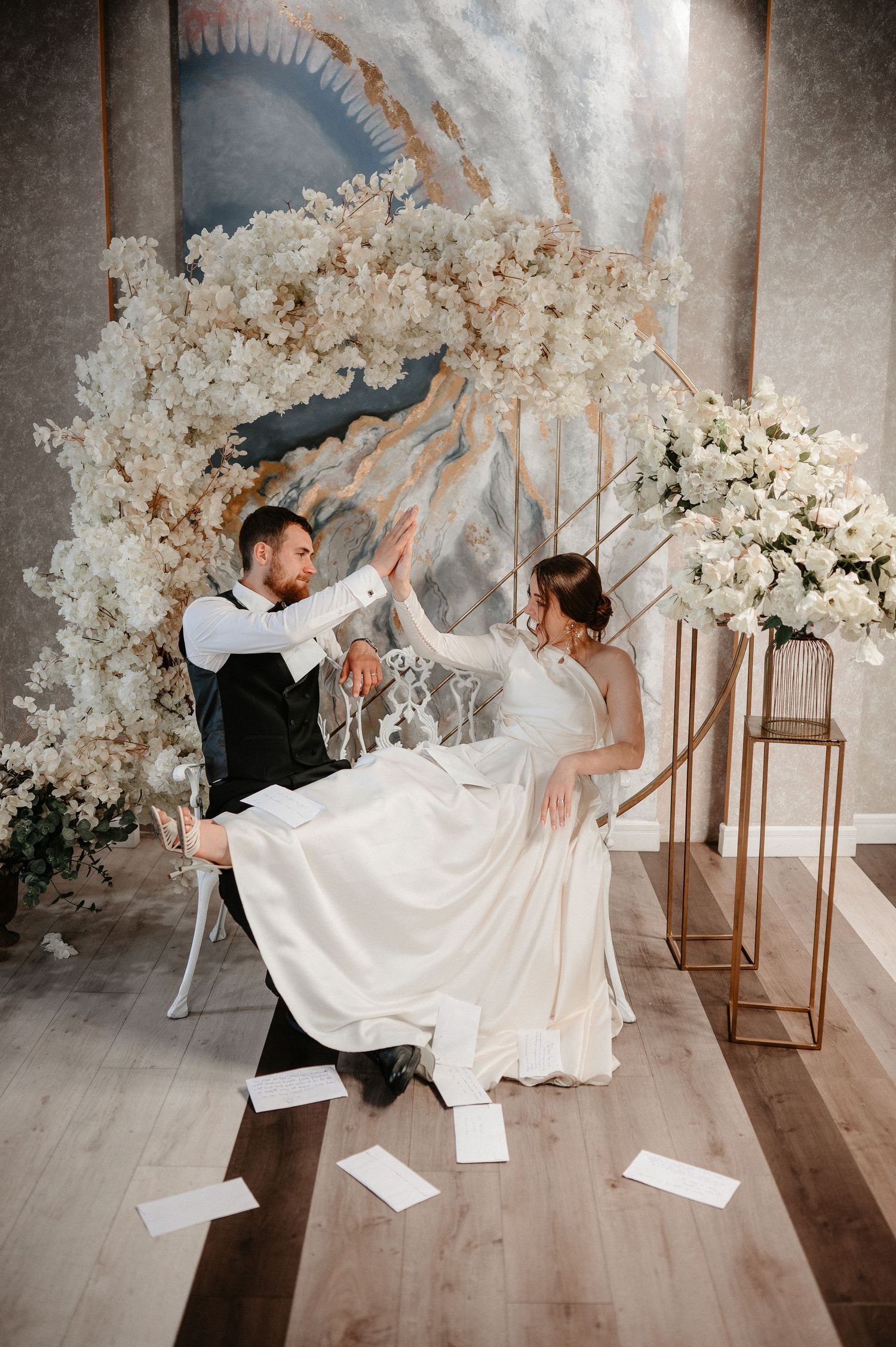 Bride and groom high-fiving joyfully on a white bench surrounded by white floral wedding decorations.