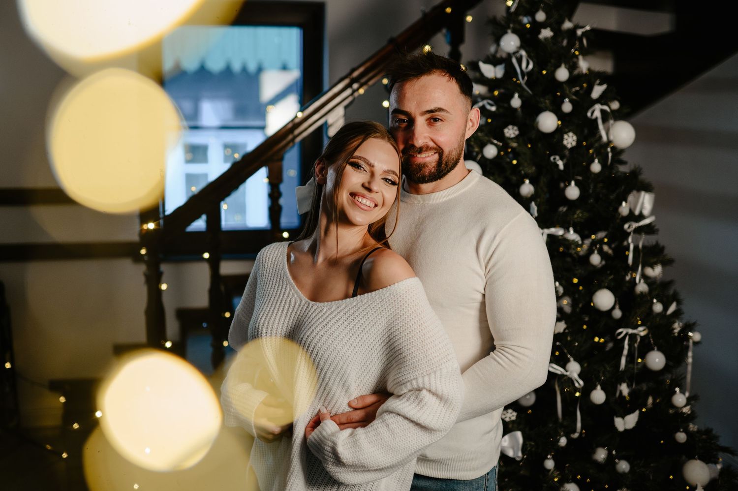 Smiling couple in white sweaters embracing near a decorated Christmas tree with bokeh lights.