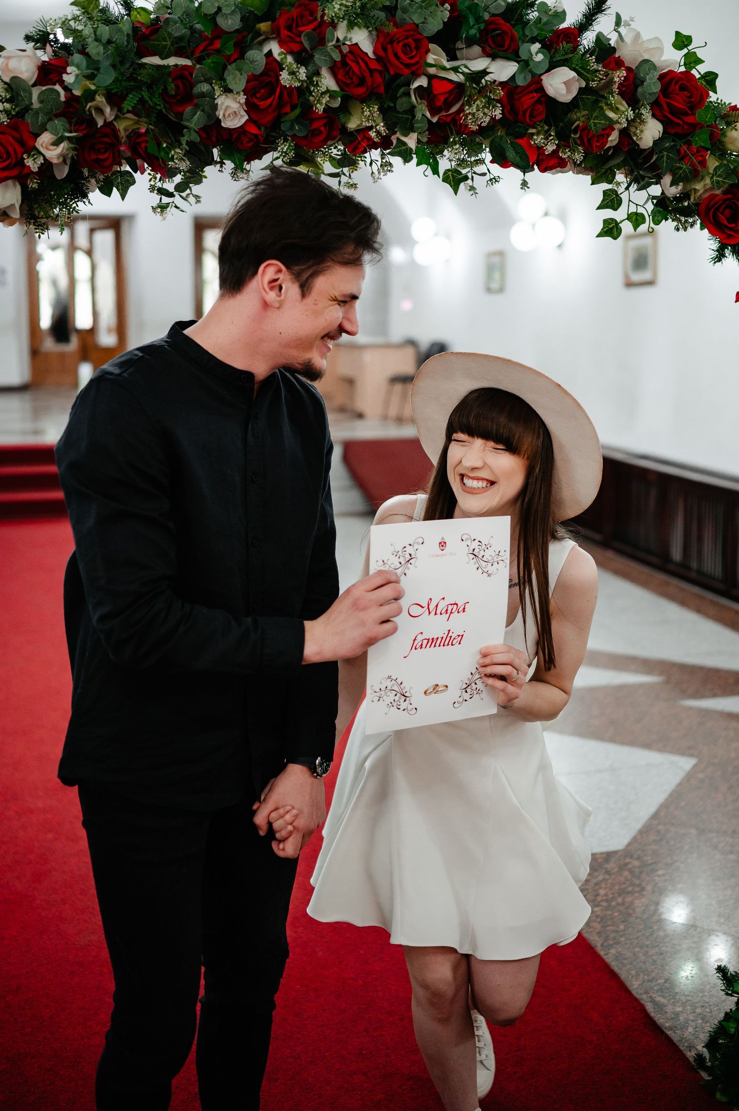 Newlyweds laugh together as bride in white dress and hat holds marriage certificate under red rose floral arch.