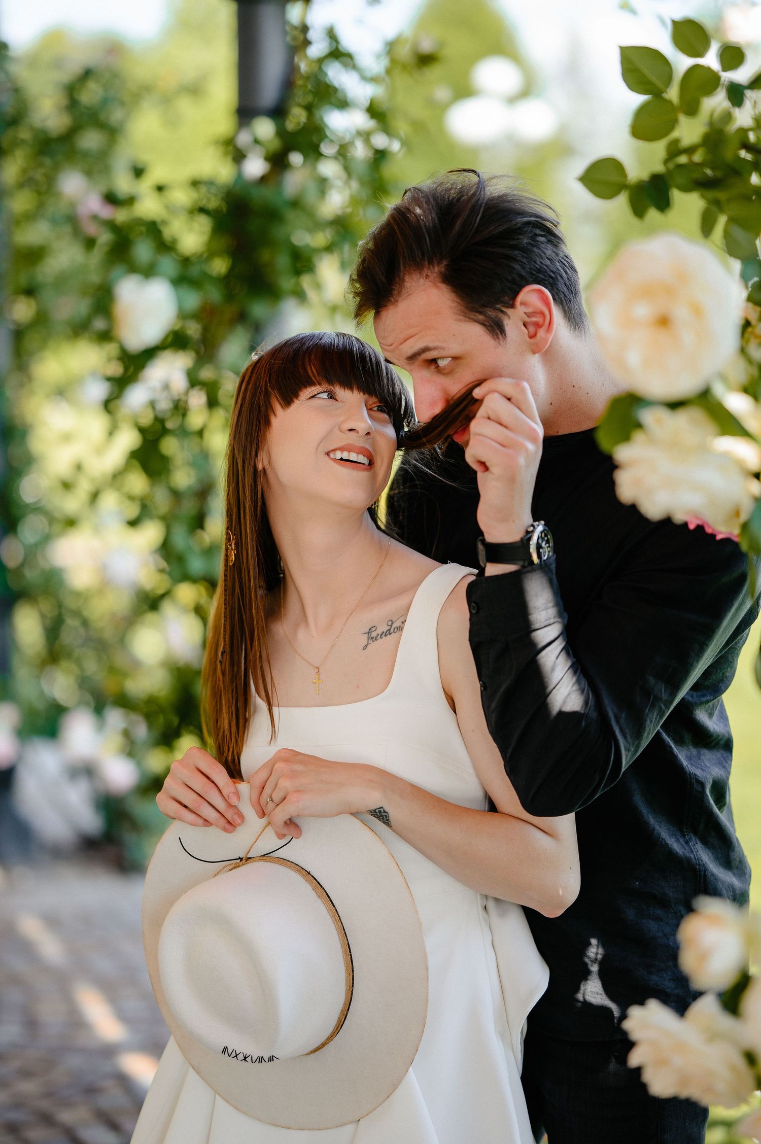 Couple smiling together among white roses, man kissing womans hand as she holds a hat.