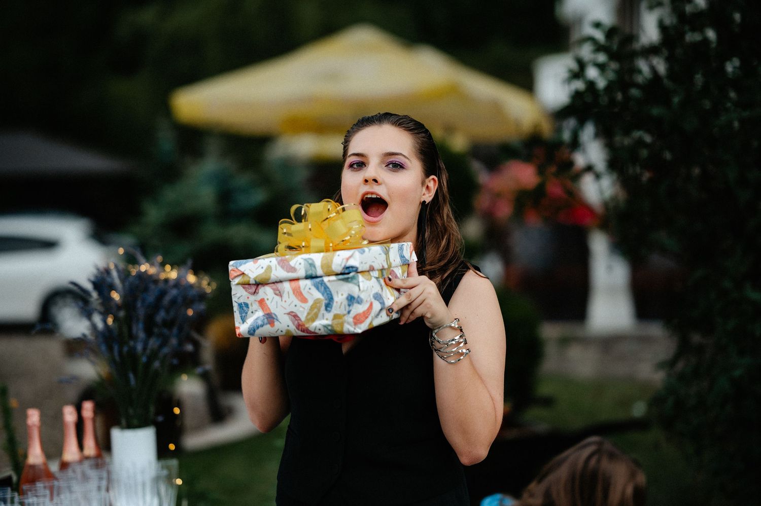 Young woman in black dress reacting with surprise while opening a colorful gift at an outdoor party.