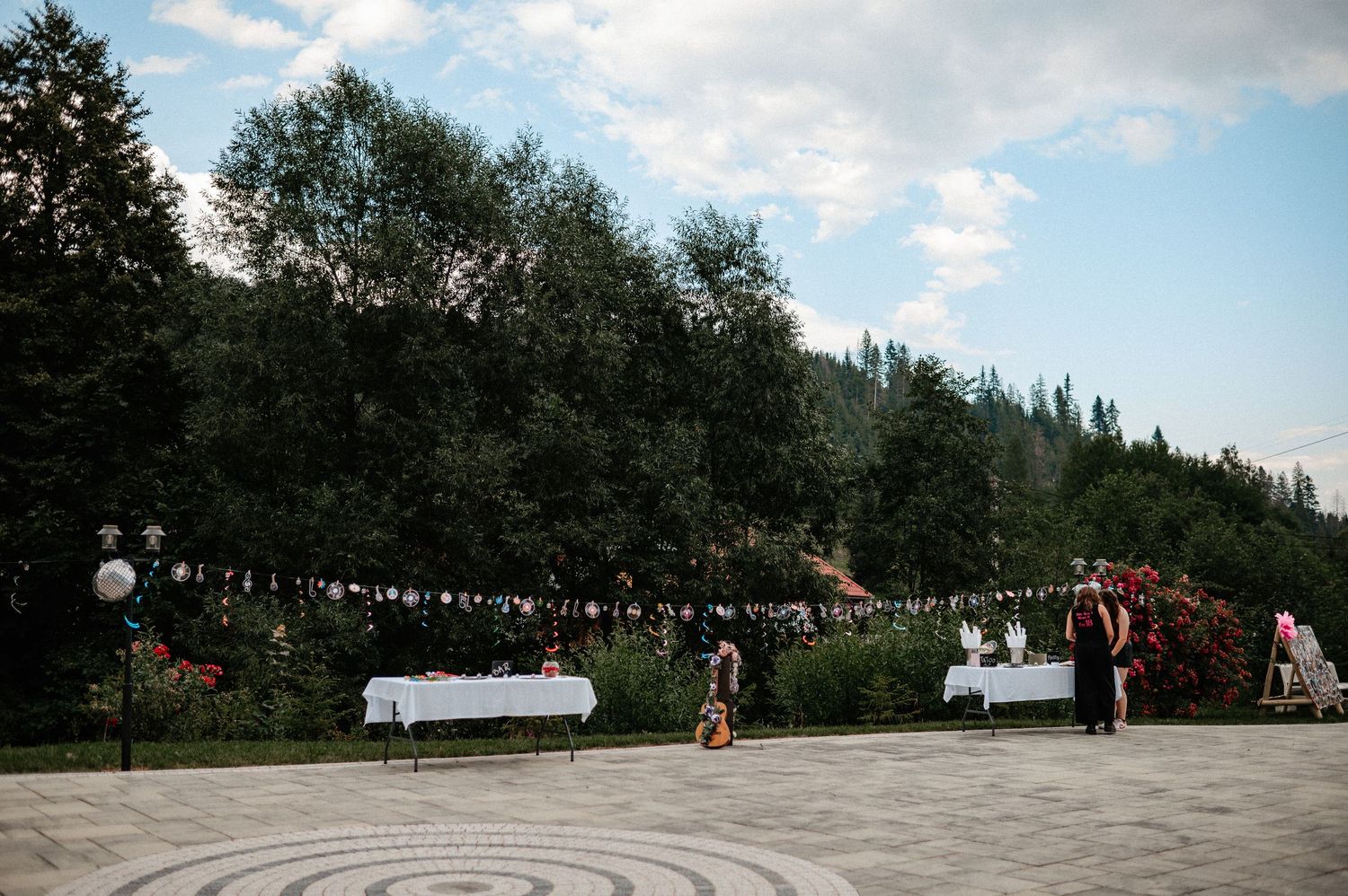 Outdoor event setup with white tables, bunting flags, and tall trees under blue sky.