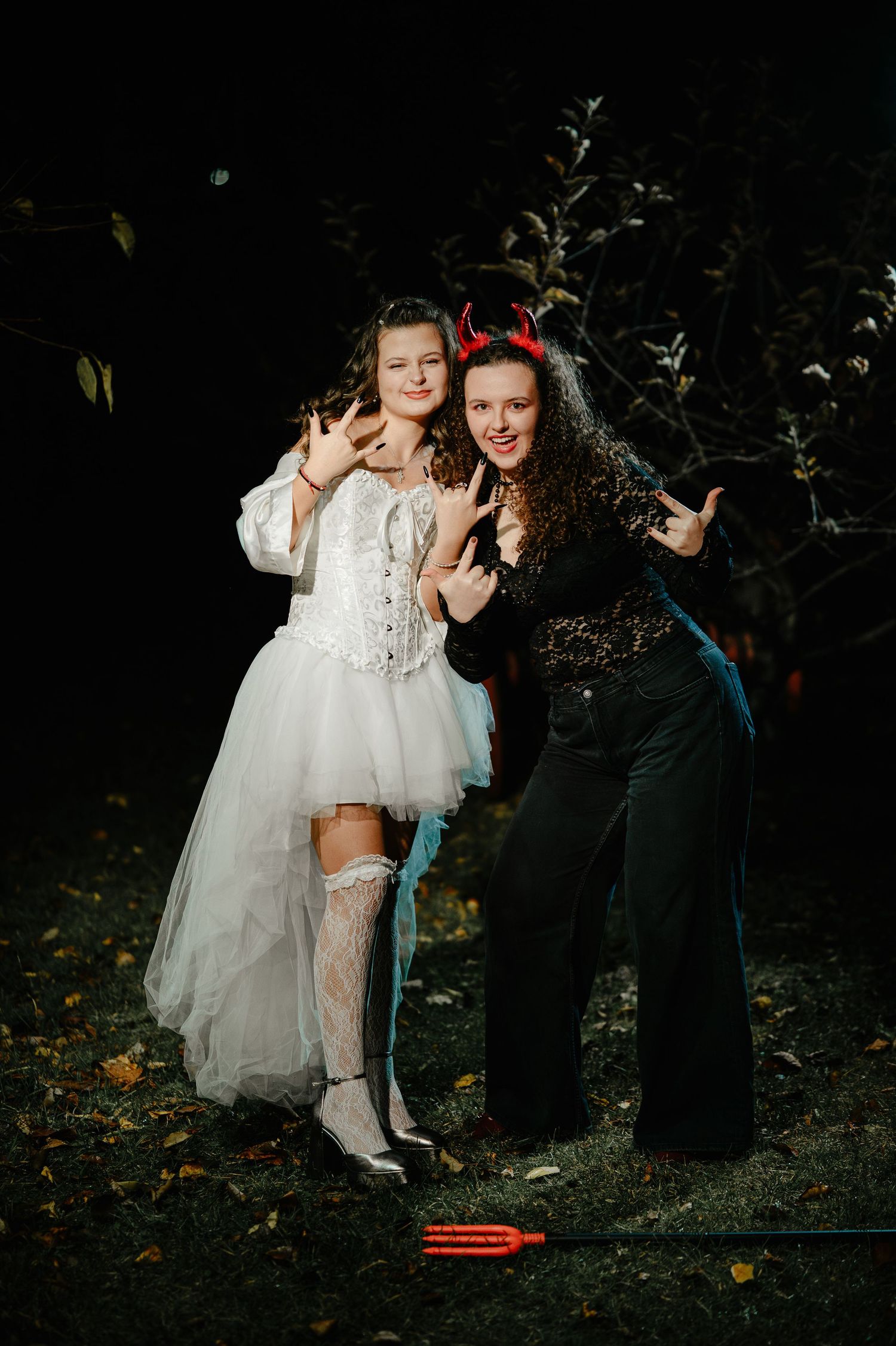 Two girls in Halloween costumes, one as angel in white corset dress, one as devil with red horns.