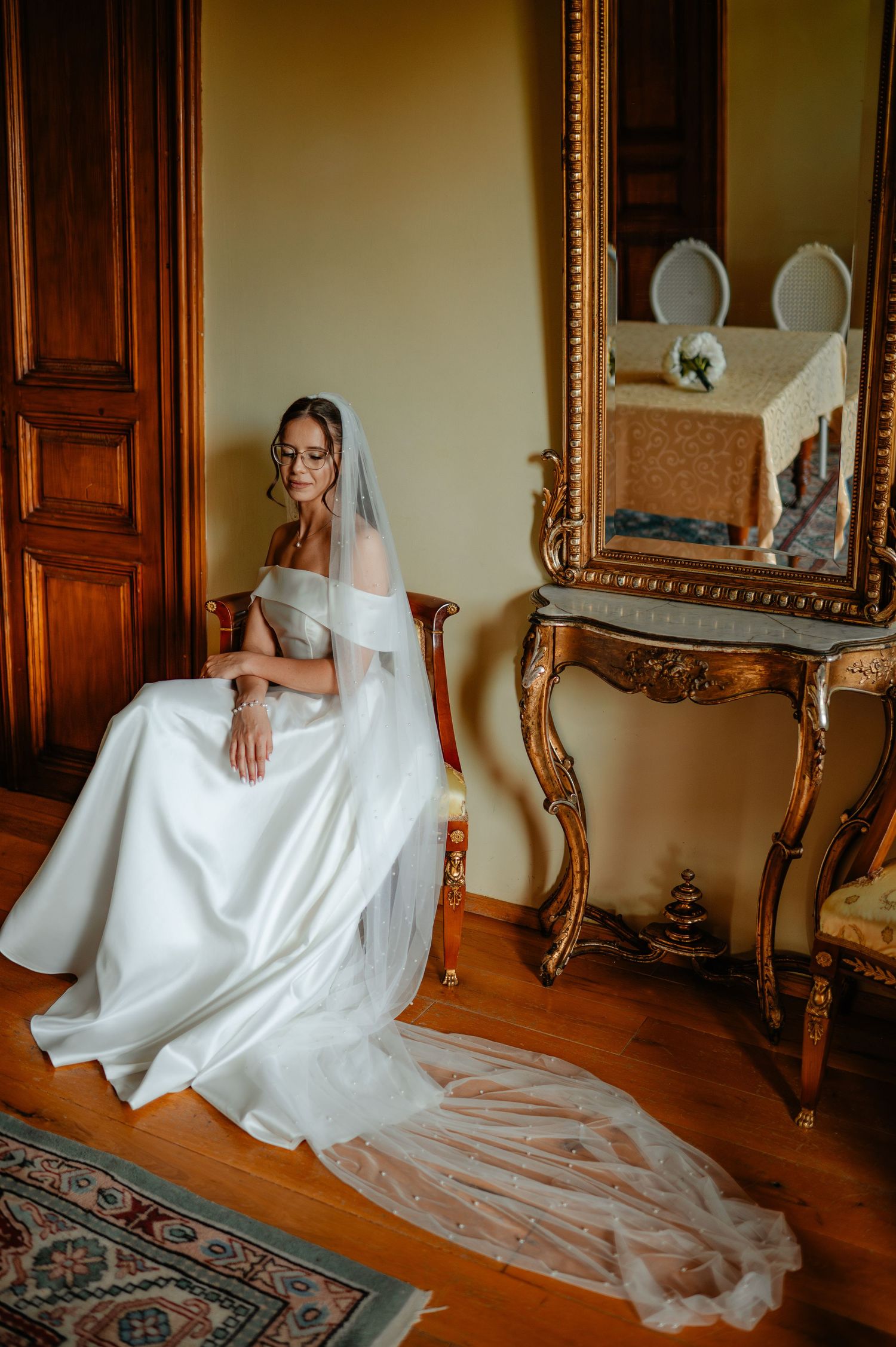 Elegant bride in off-shoulder white gown and veil seated beside ornate gold mirror in vintage room.