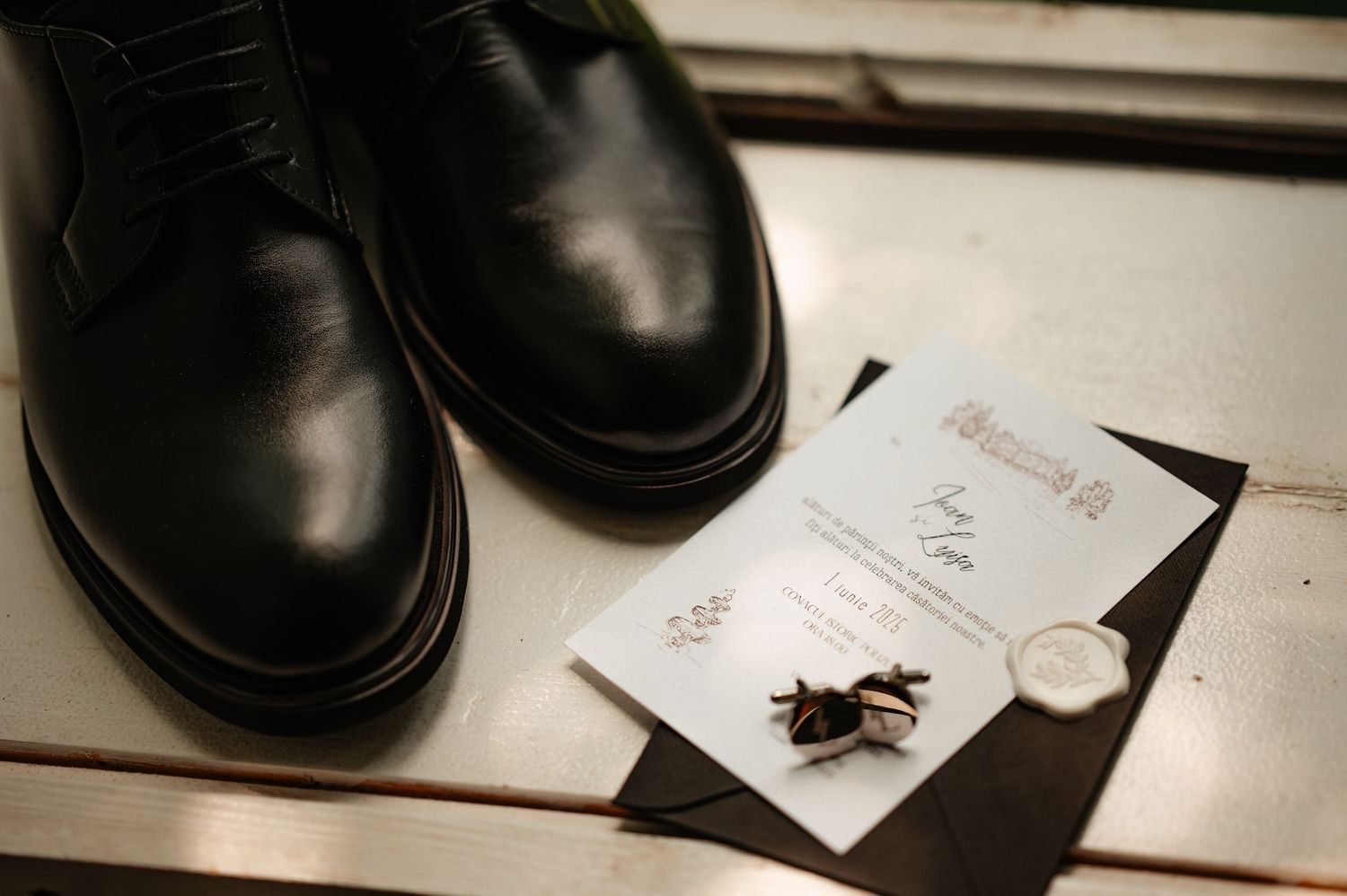 Black leather Oxford groom shoes with wedding invitation, wax seal, and cufflinks on white surface.