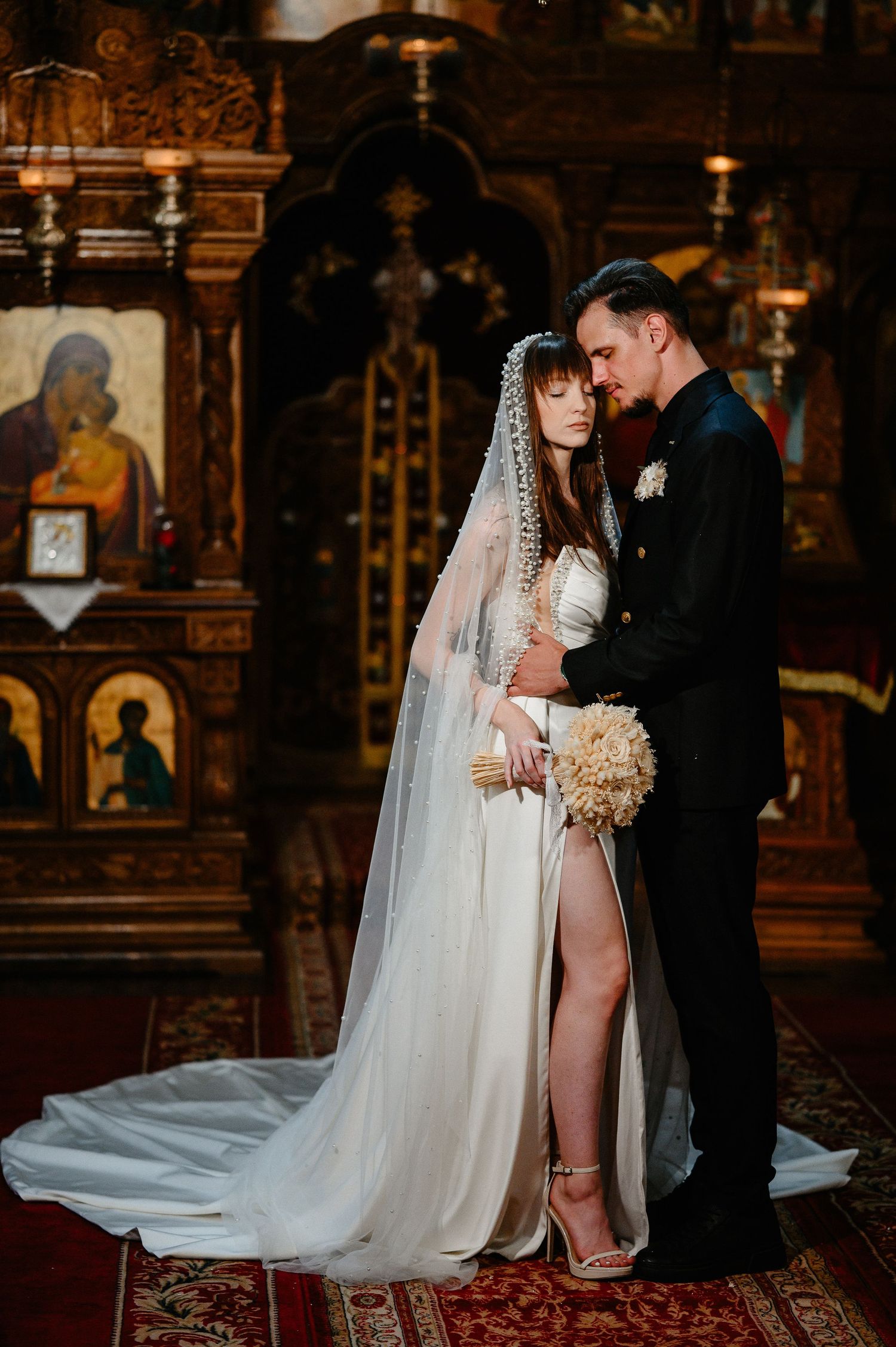 Bride and groom embrace in Orthodox church interior with golden iconostasis during wedding ceremony.