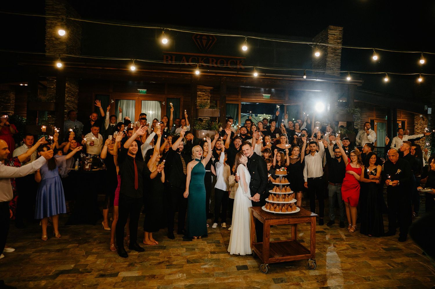 Large wedding crowd raises glasses in toast as couple cuts cake at outdoor evening reception.