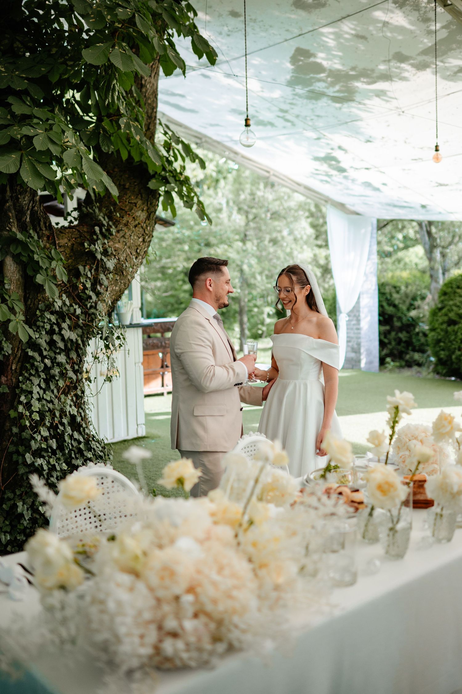 Bride in white off-shoulder mini dress and groom in beige suit share a laugh beside floral table arrangements.