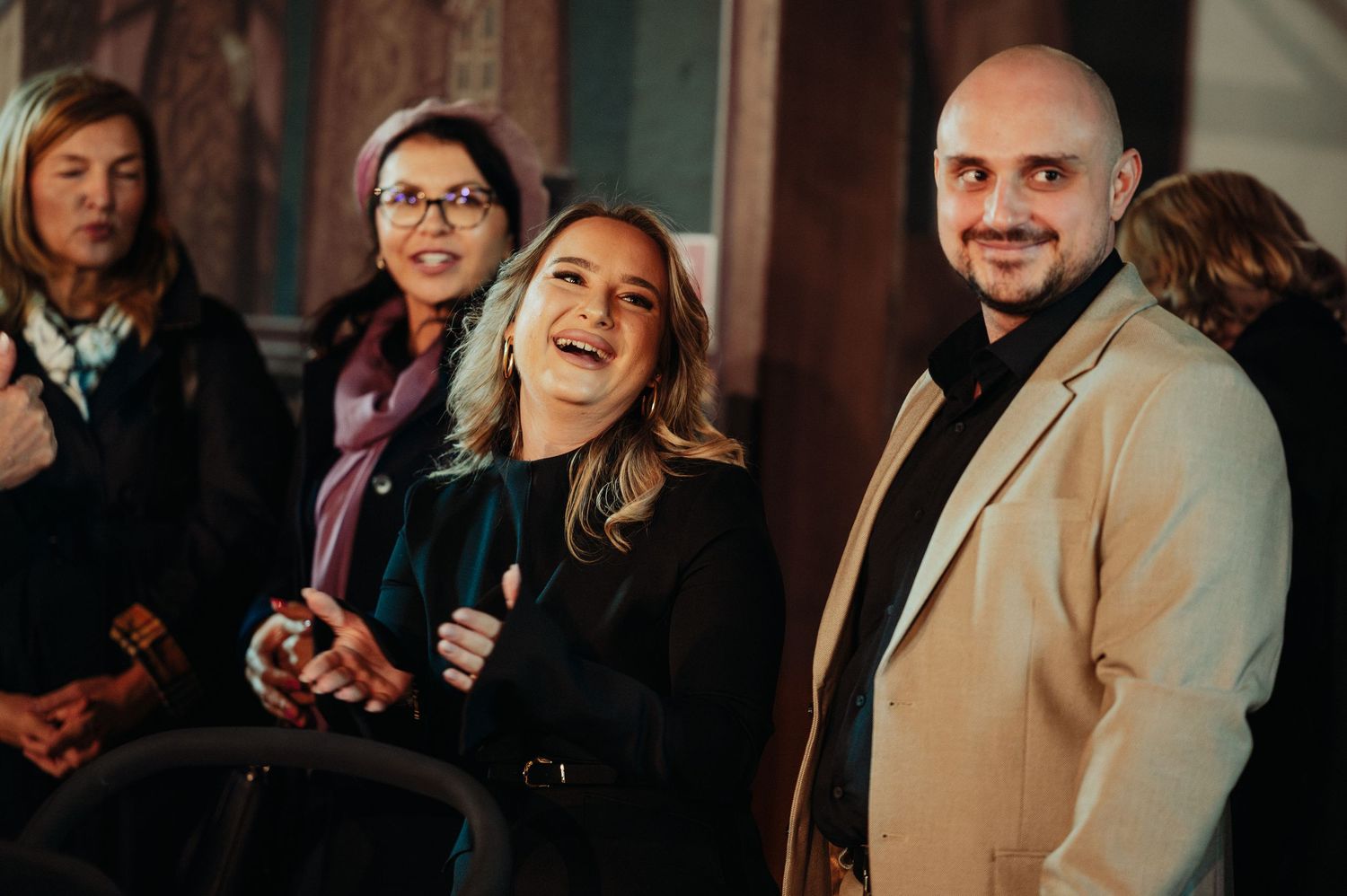 Group of smiling adults at an indoor event, with a laughing woman in a black jacket.