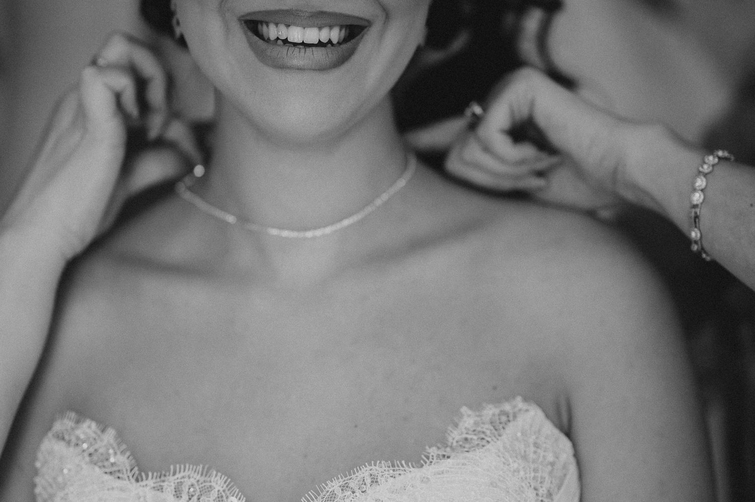 Black and white close-up of smiling bride in lace wedding dress having pearl earring put on.