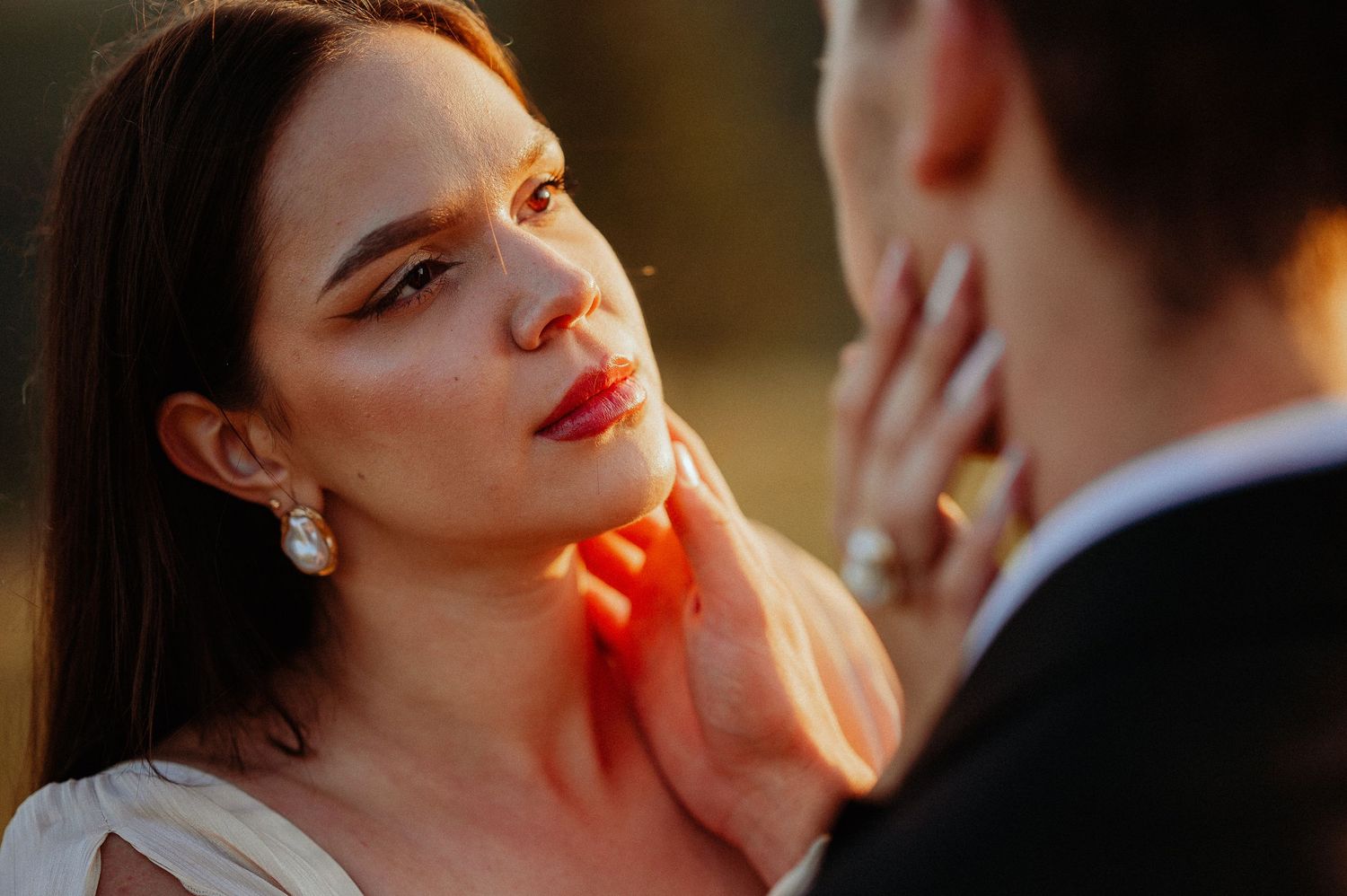 Close-up of bride with red lips and pearl earring gazing at groom in warm golden light.