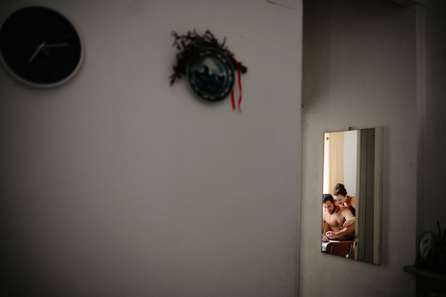 Couple embracing reflected in a mirror on a dimly lit room wall with a decorative clock nearby.