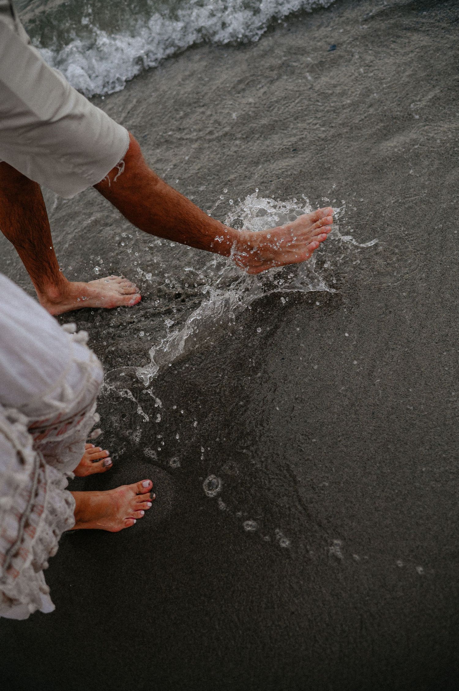 Close-up of couple splashing bare feet in ocean waves on a sandy beach.