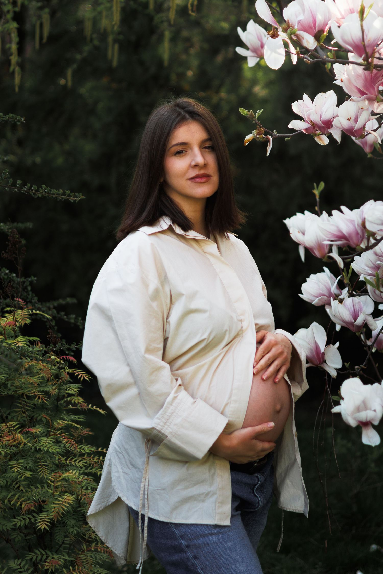 Pregnant brunette woman in cream shirt holding belly beside blooming pink magnolia flowers outdoors.