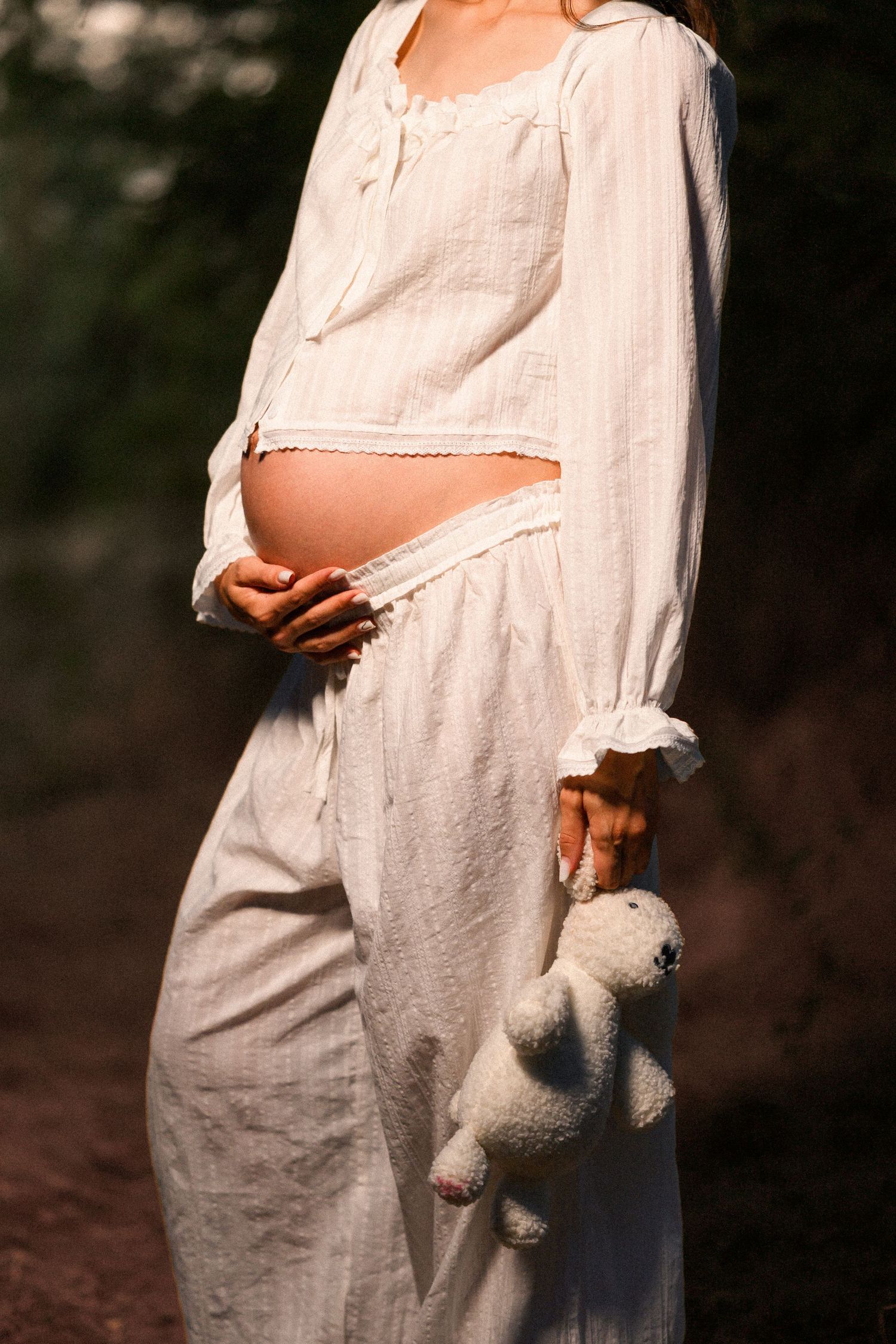 Pregnant woman in white linen outfit holding a teddy bear, cradling her baby bump outdoors in soft natural light.