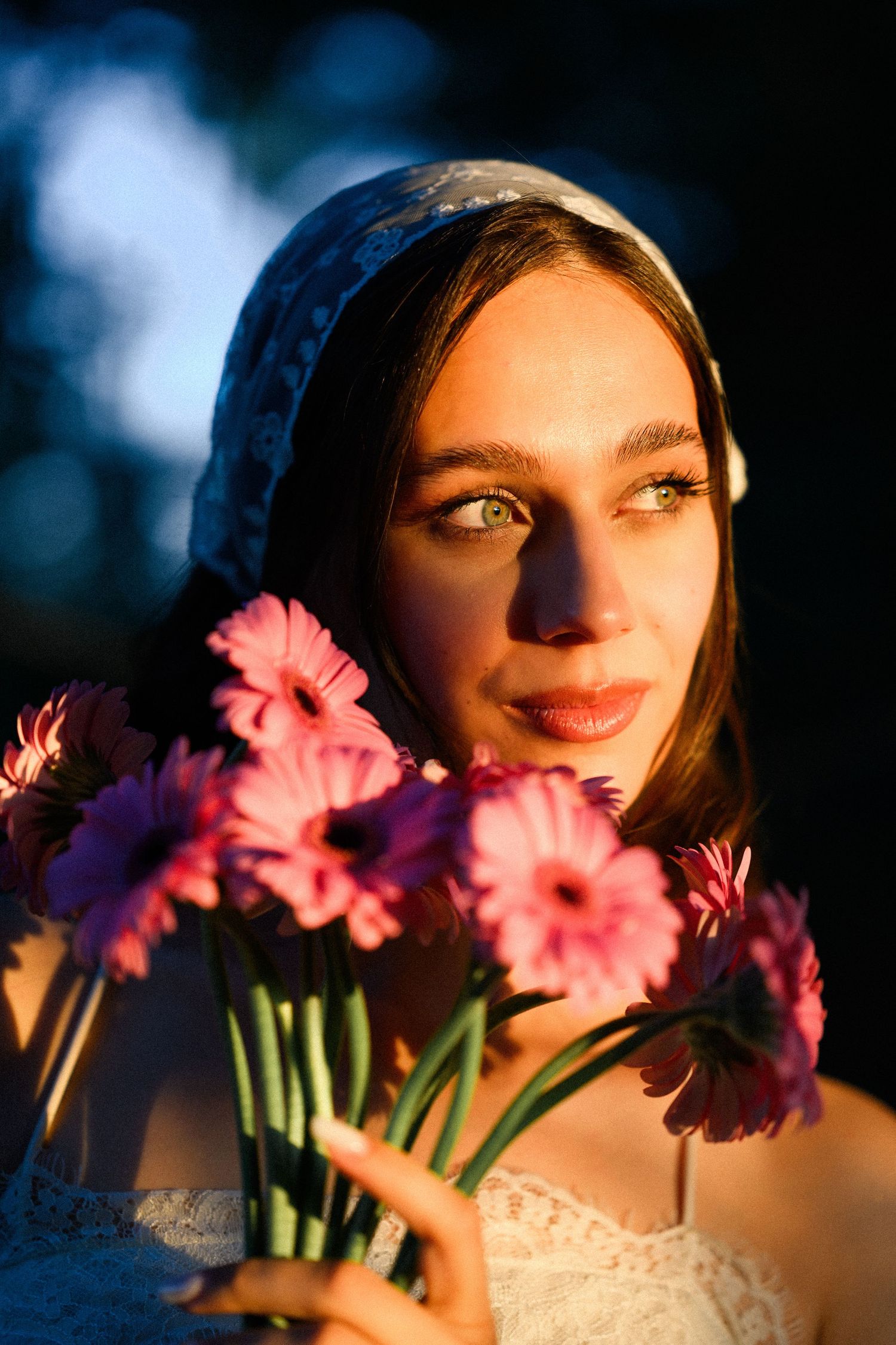 Young woman with blue headscarf holding pink gerbera daisies in golden hour sunlight, gazing thoughtfully into distance.