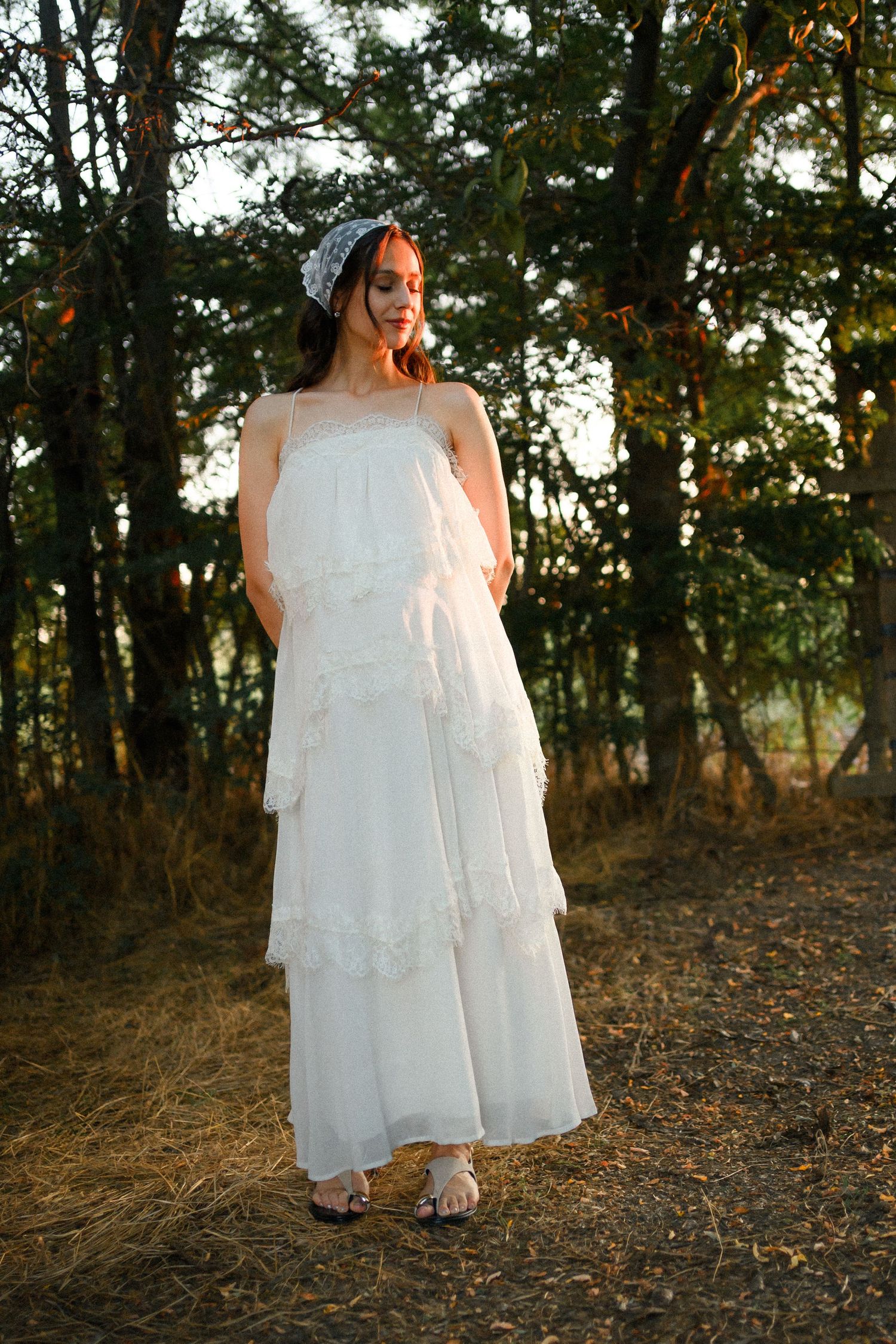 Woman in white tiered maxi dress and headscarf standing in a sunlit forest clearing at golden hour.