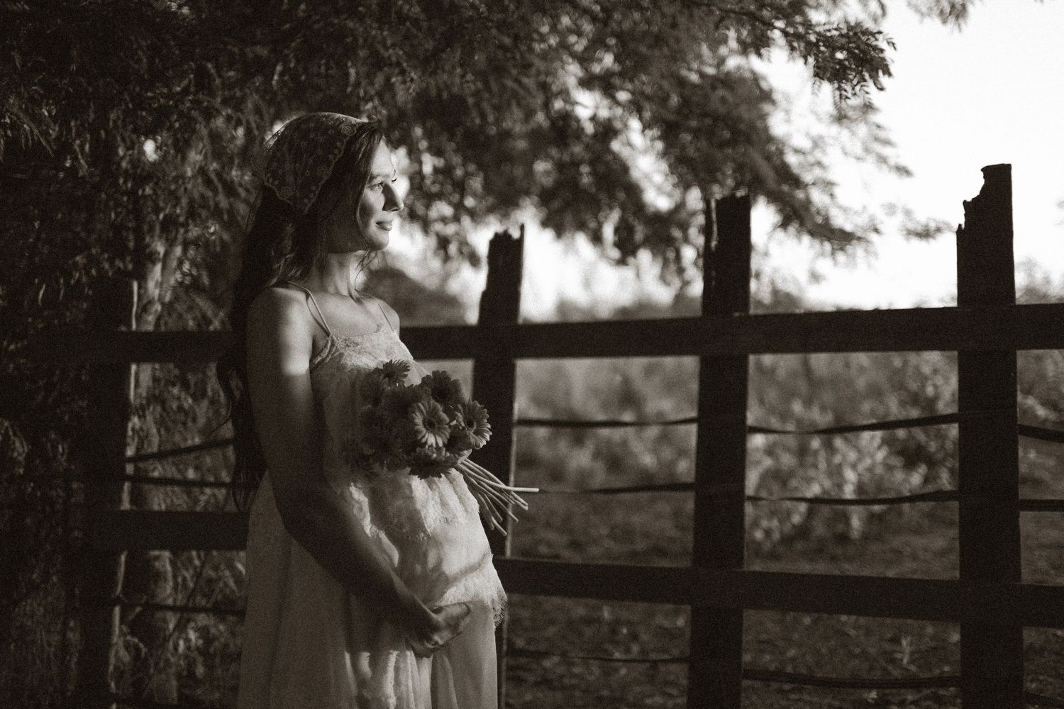 Pregnant woman in white dress holding flowers by rustic fence in black and white maternity photo.
