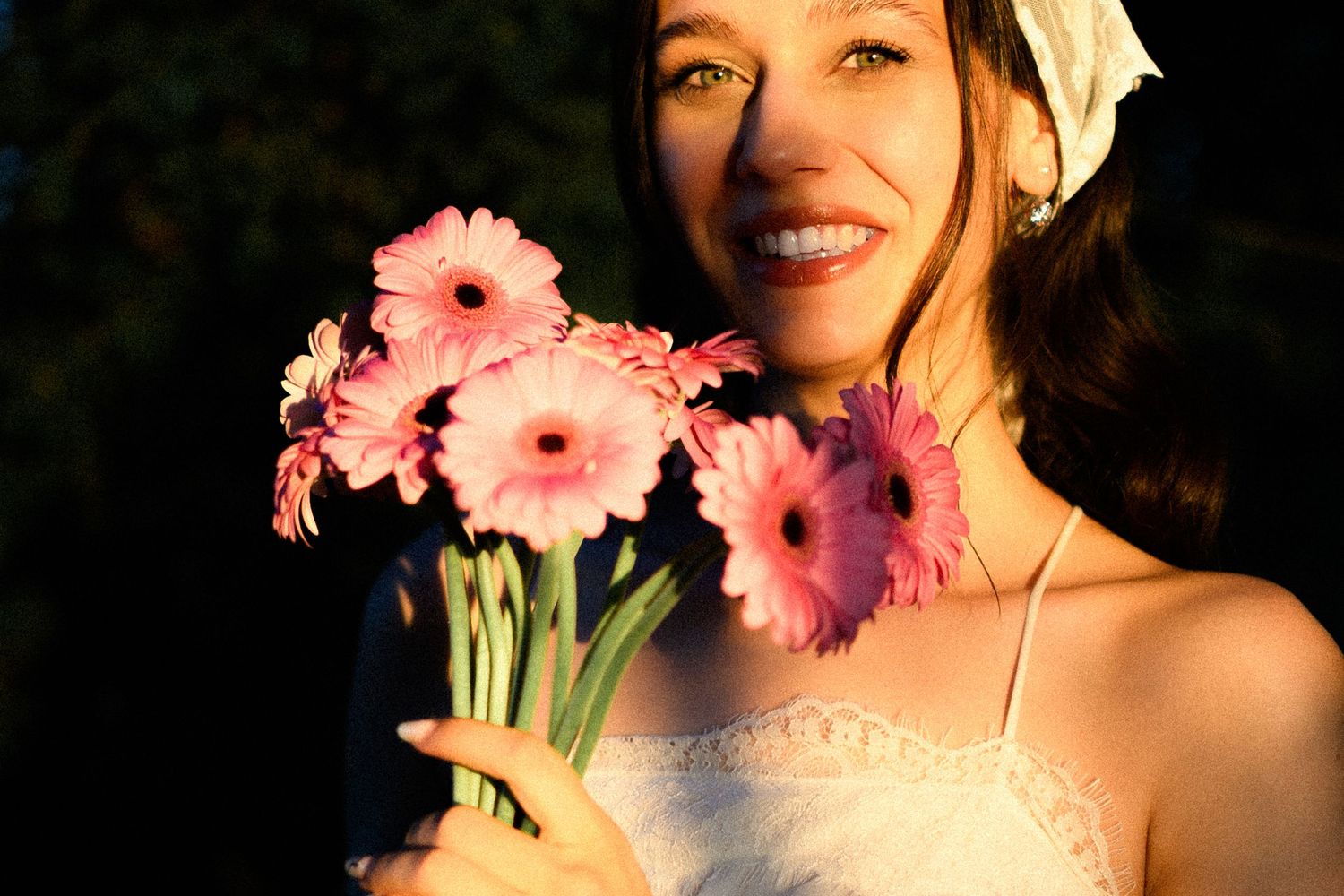 Smiling young woman holding a bouquet of pink gerbera daisies outdoors in warm golden sunlight.