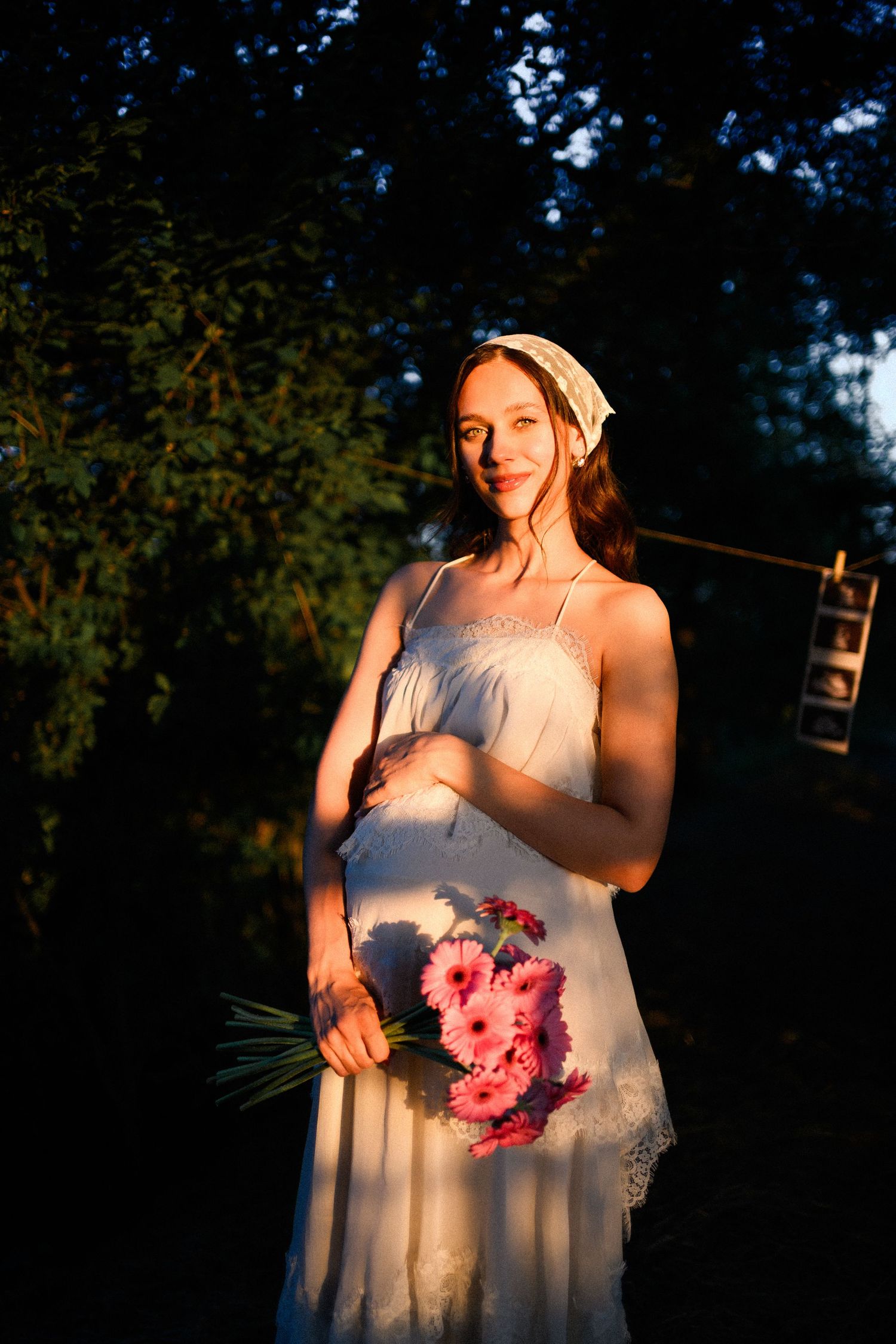 Pregnant woman in white sundress holding pink gerbera bouquet, bathed in golden sunset light among green trees.