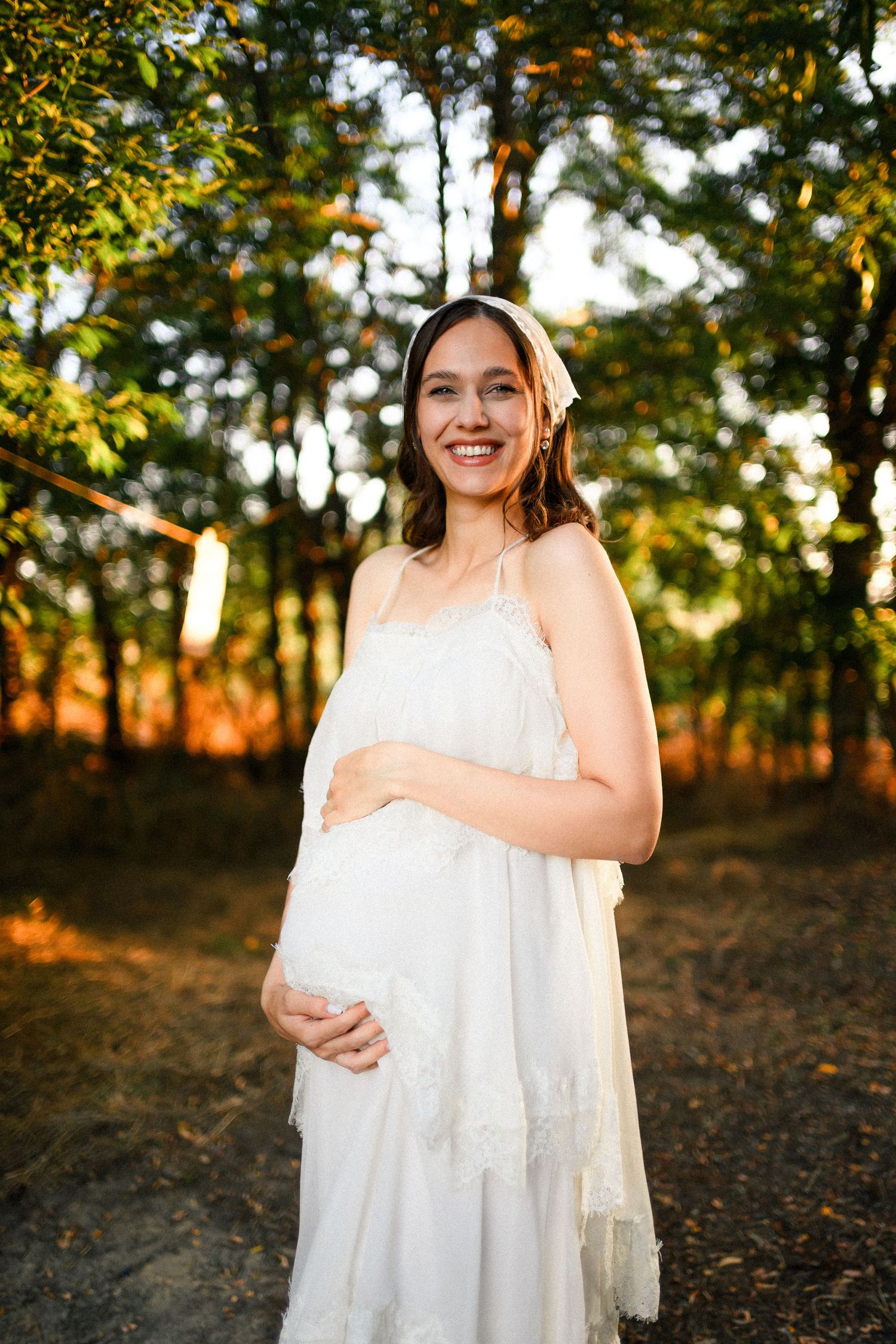 Smiling pregnant woman in white lace dress holding belly, standing in sunlit forest during golden hour.