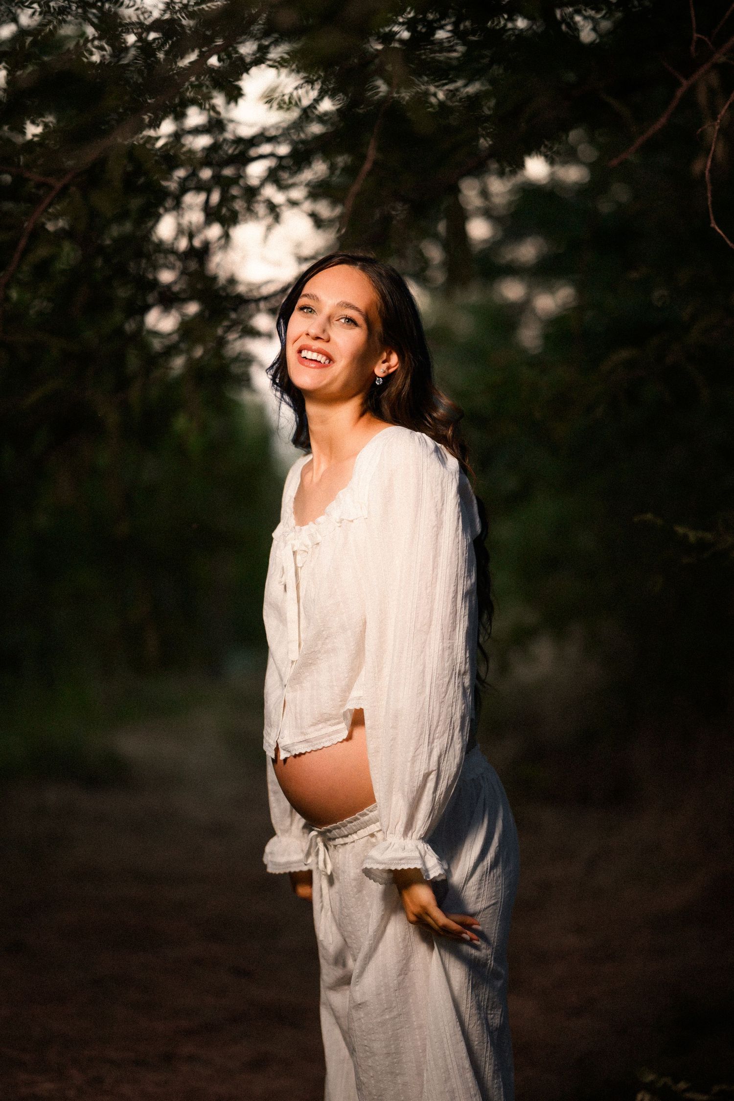 Smiling pregnant woman in white linen outfit standing in a sunlit forest path, cradling her baby bump.