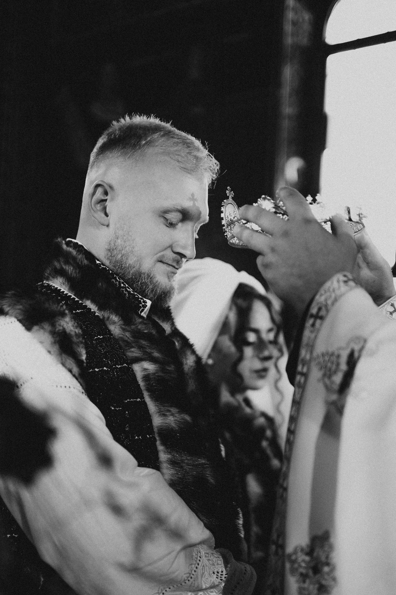 Black and white photo of a groom receiving a crown blessing from a priest during an Orthodox wedding ceremony.