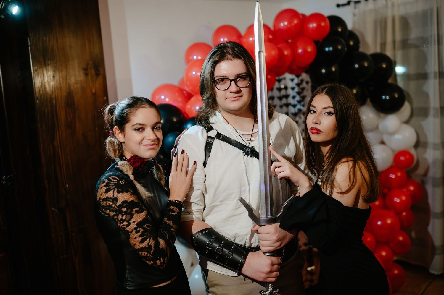 Three people in Halloween costumes pose with a large sword in front of red and black balloons.