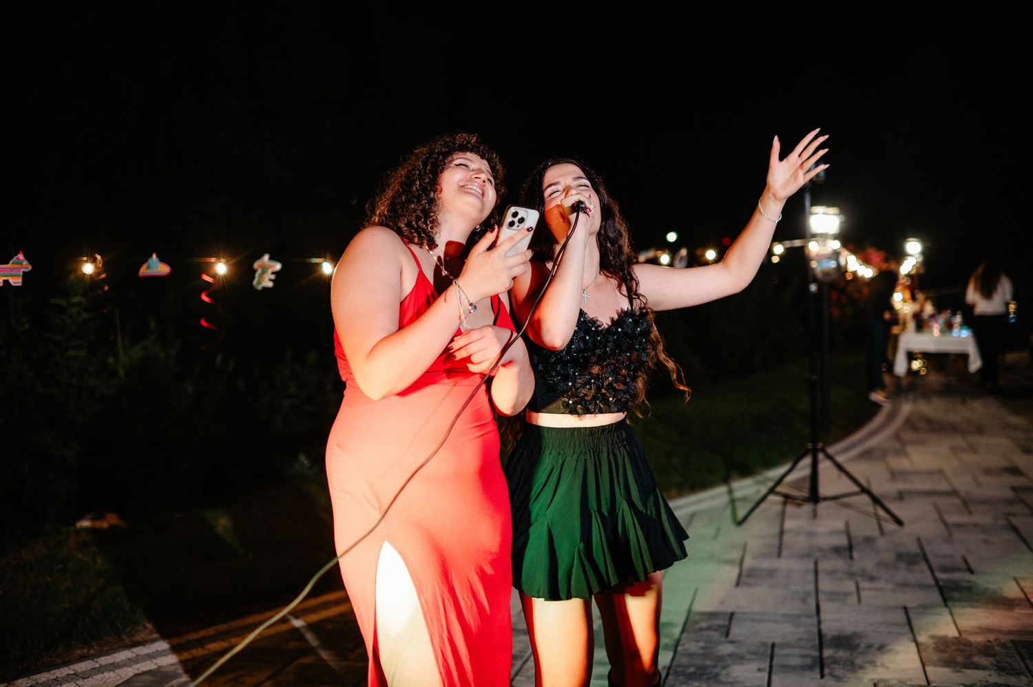 Two women singing on stage at night outdoor event, one in red dress and one in black and green outfit.