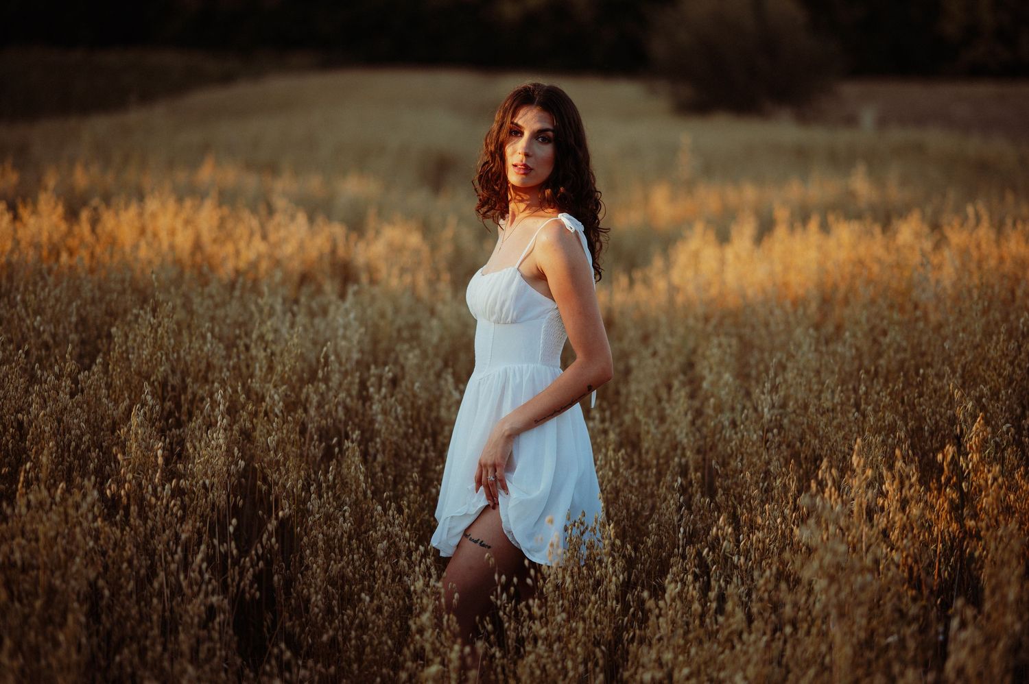Elegant woman in white dress standing gracefully in a golden wheat field at sunset.