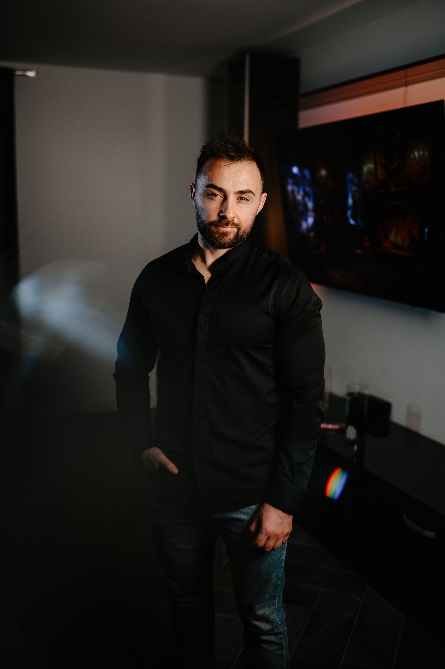 Confident bearded man in black shirt standing in dark modern room with TV.