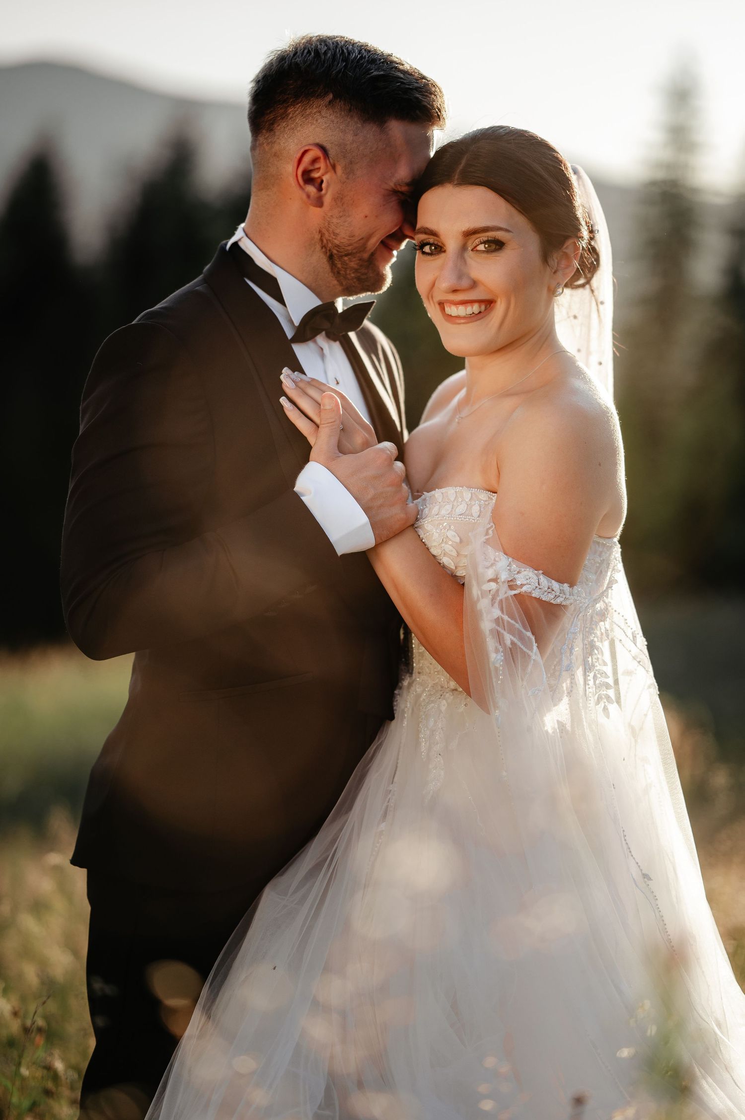 Smiling bride in an off-shoulder tulle gown embraces groom in golden hour mountain wedding portrait.