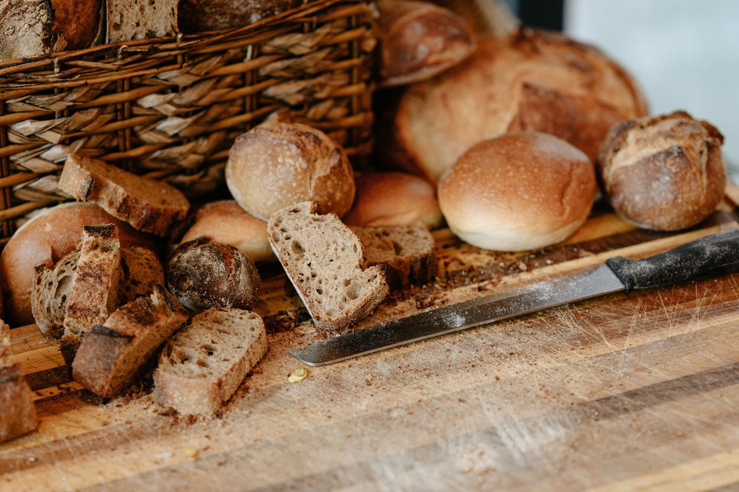 Assorted rustic artisan breads and rolls scattered on a wooden cutting board with a bread knife.