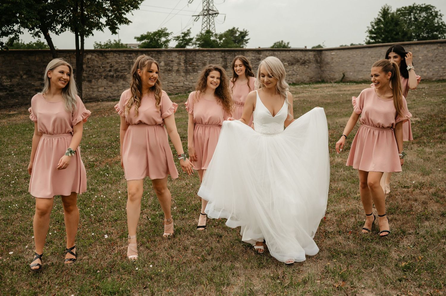 Bride in a flowing white gown walks and laughs with bridesmaids in pink dresses on a lawn.