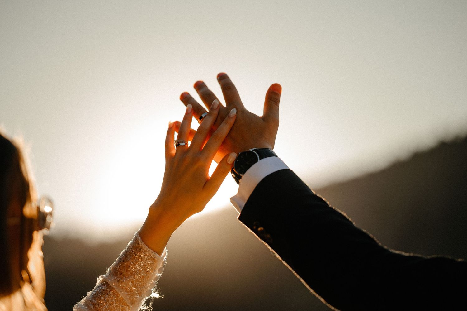 Close-up of bride and groom touching hands against golden sunset sky on wedding day.