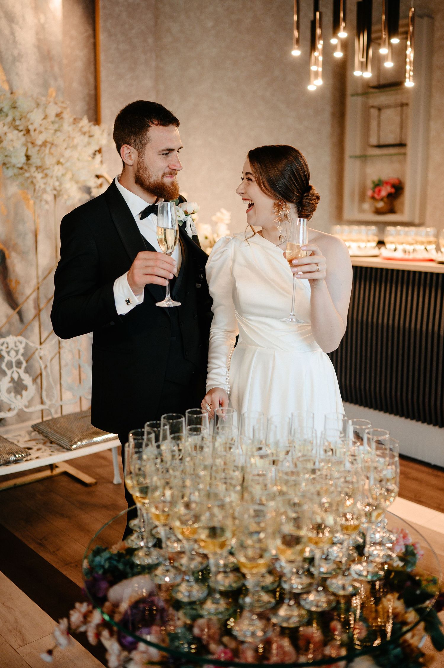 Bride and groom toasting champagne beside pyramid of glasses at wedding reception.