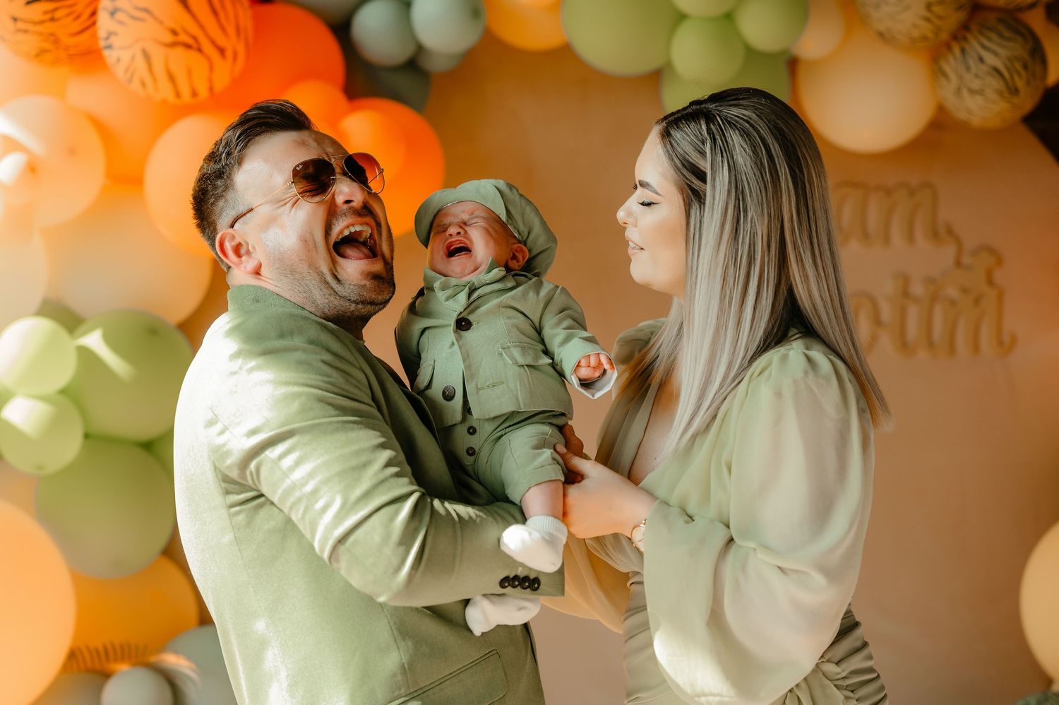 Laughing father in green suit holding crying newborn while mother smiles at balloon party backdrop.