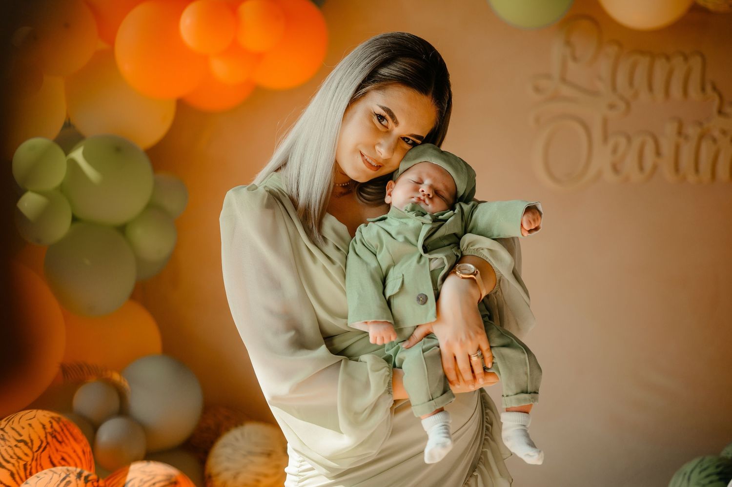 Young mother in sage green outfit holding sleeping newborn at orange balloon celebration backdrop.