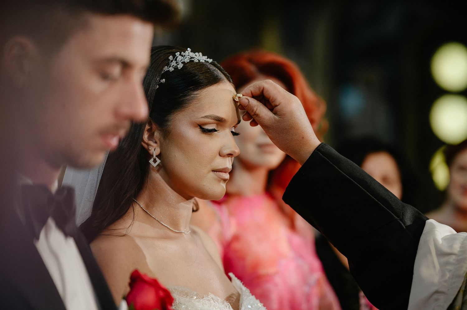 Bride wearing a crystal tiara being anointed during an Orthodox wedding ceremony with guests watching.