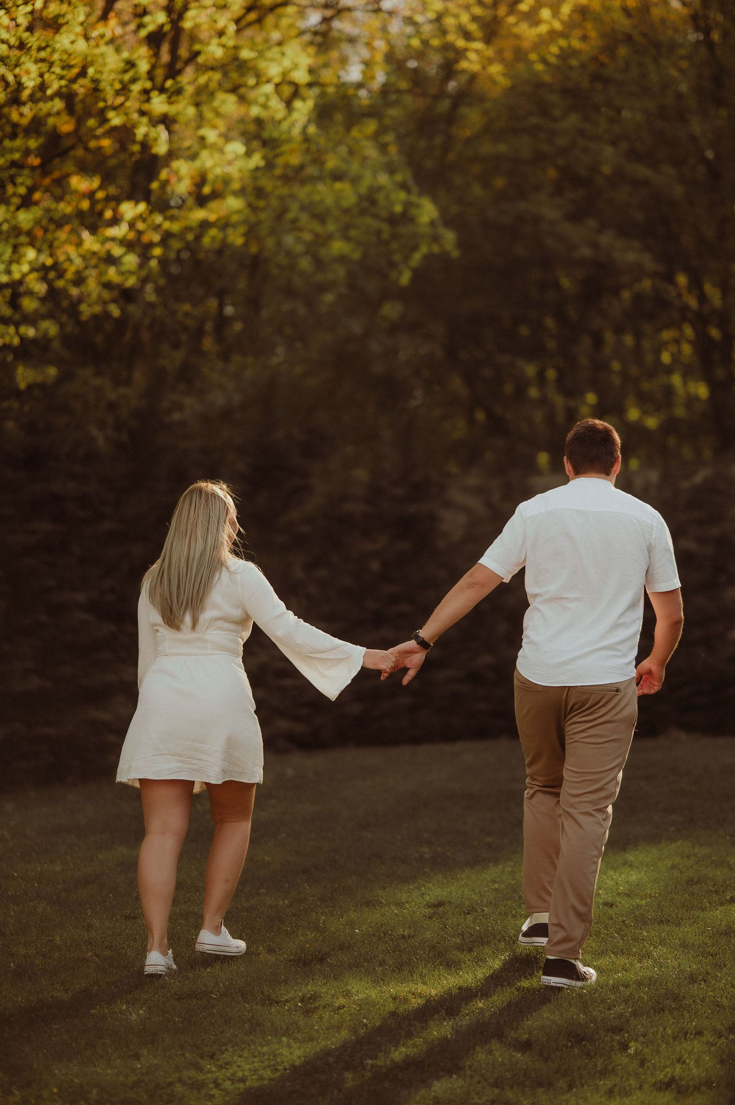 Couple holding hands walking away through lush green grass in beautiful golden hour sunlight.
