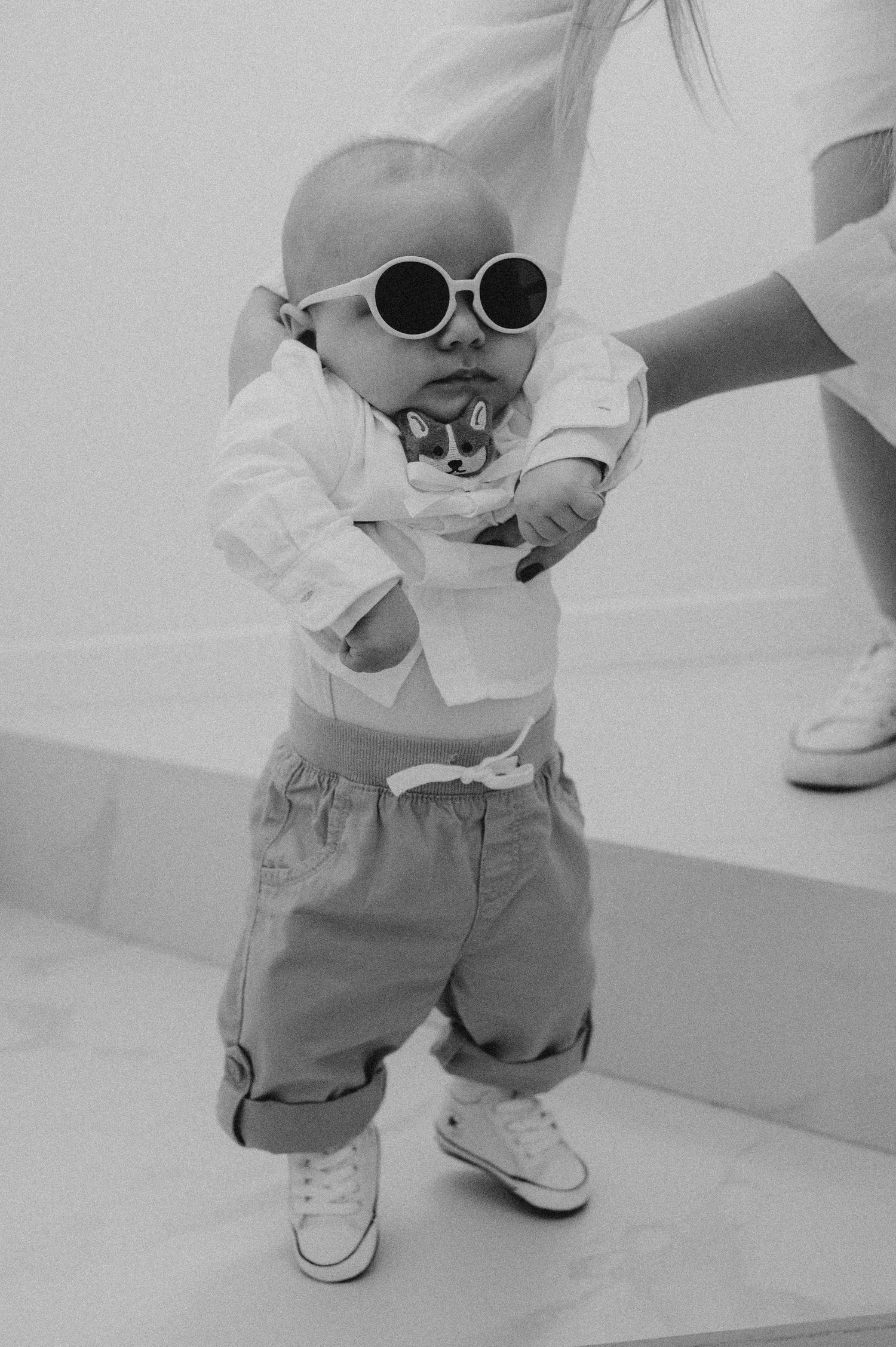 Cute baby in round sunglasses and white shirt taking first steps in black and white photo.