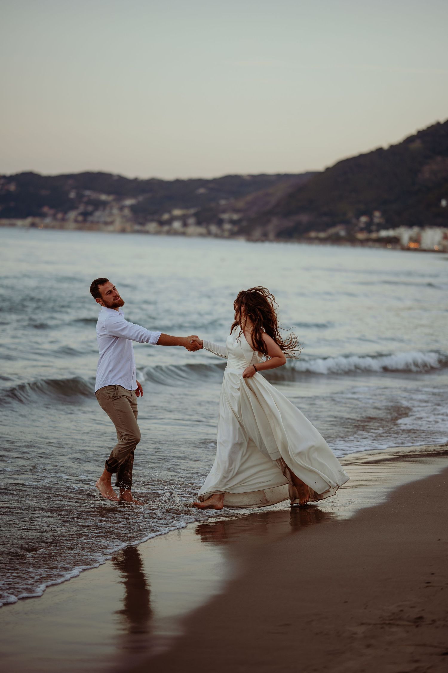 Bride and groom dancing joyfully on a beach at sunset with ocean waves.