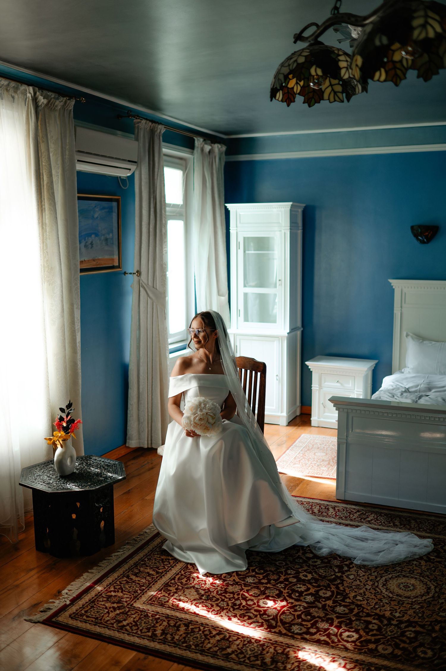 Smiling bride in white off-shoulder gown holding bouquet in blue decorated room.