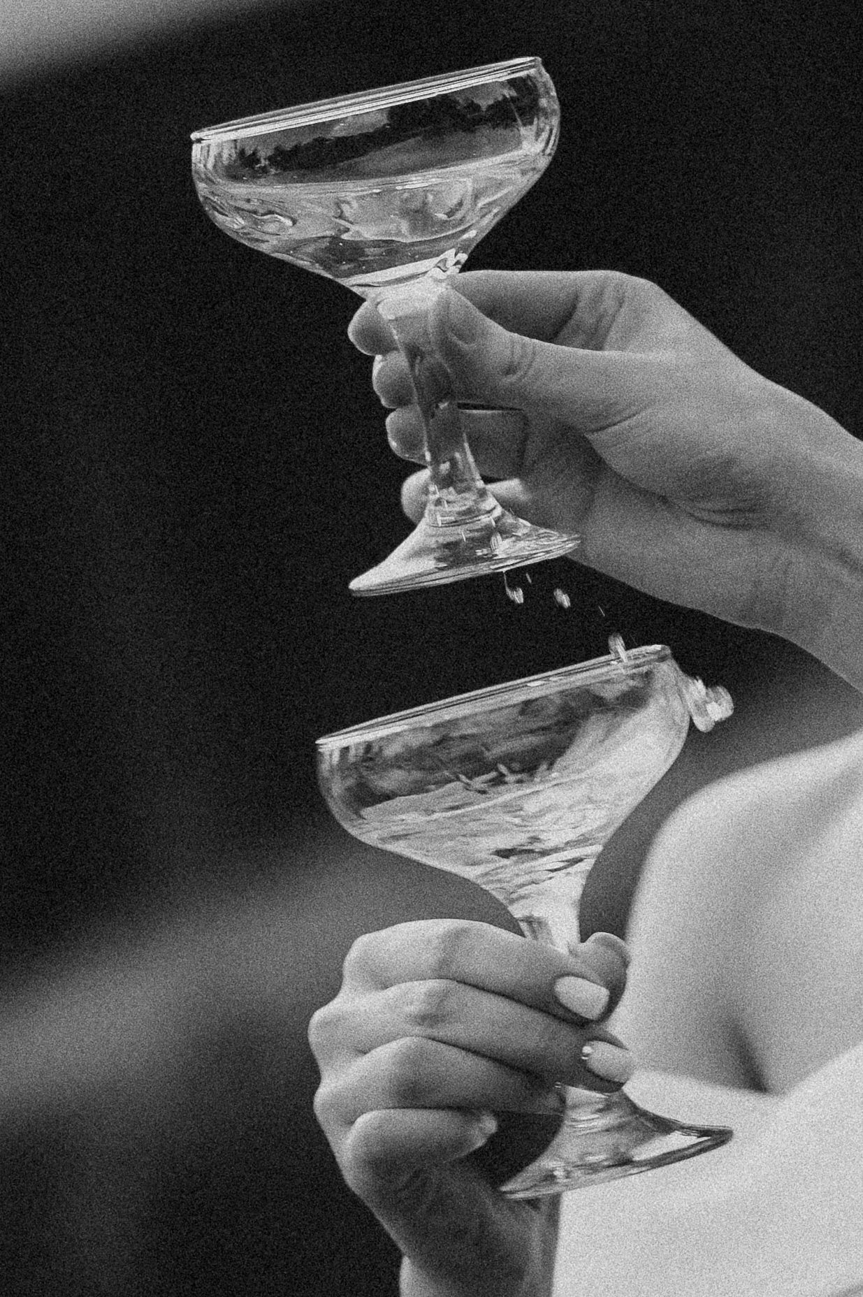 Black and white close-up of two hands clinking crystal champagne coupe glasses at a wedding toast.